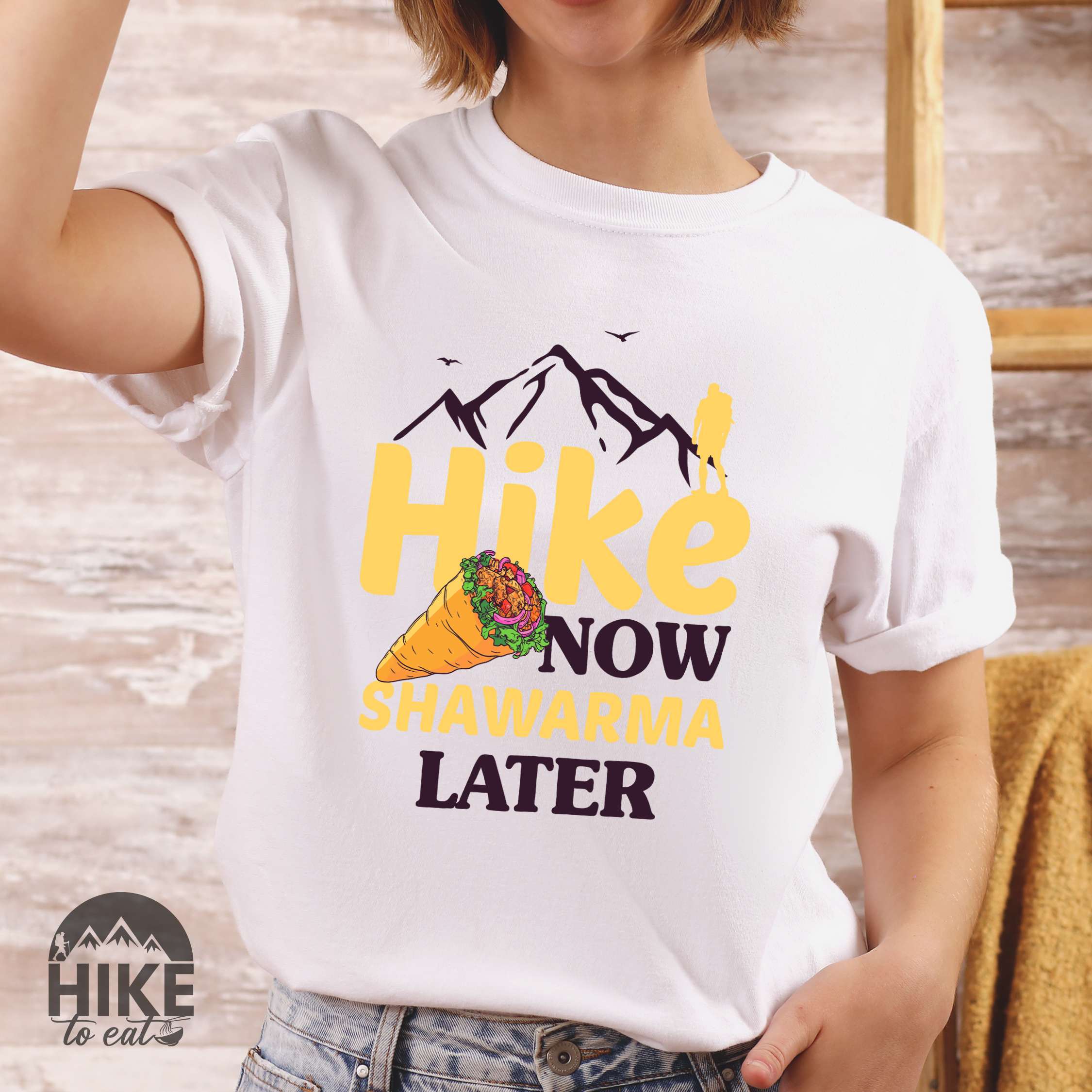 Woman wearing white "Hike now Shawarma later" colored vintage tee with bold typography, mountains, birds, and illustrated shawarma.