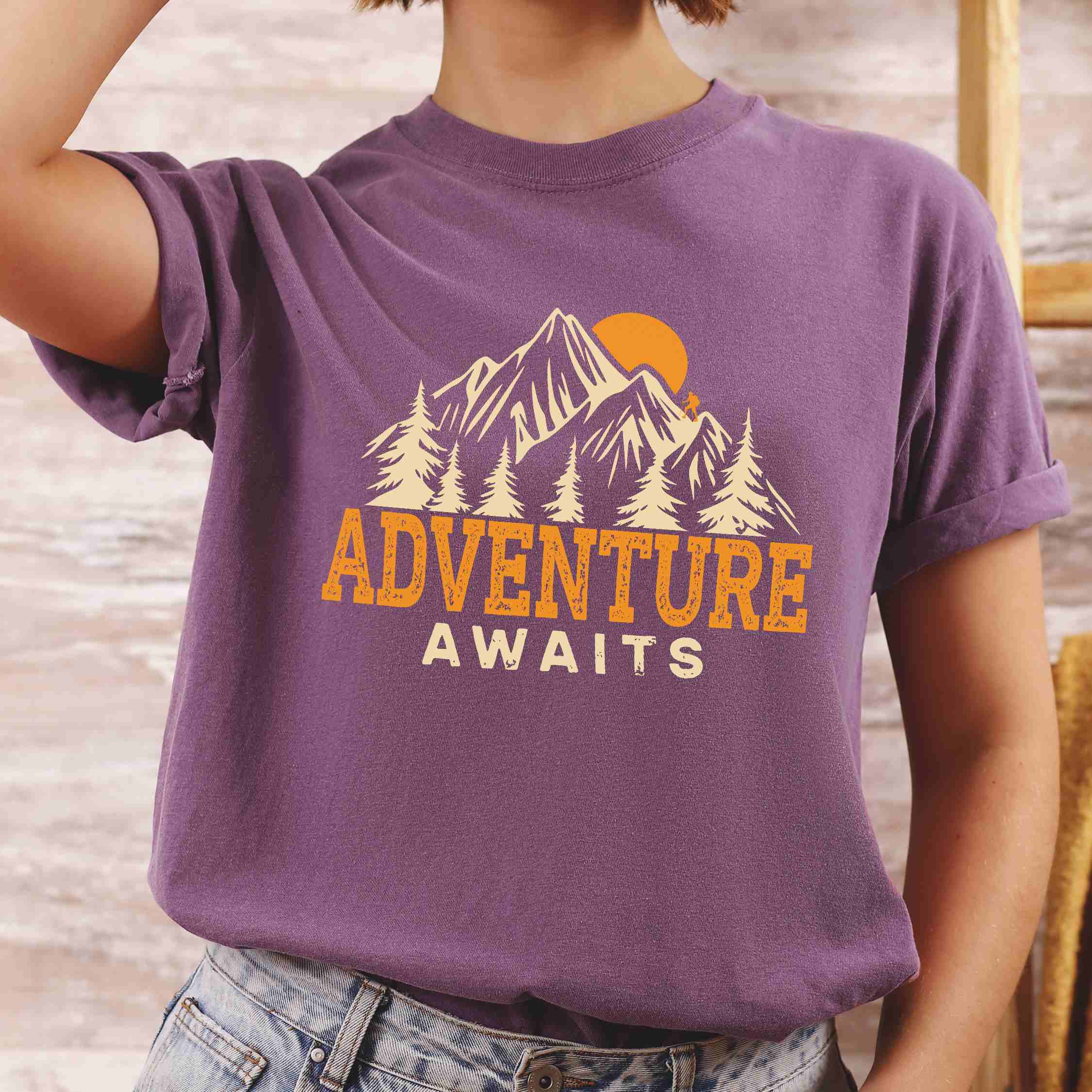 woman wearing berry "Adventure Awaits" colored statement tee with bold typography, mountains, birds, perfect for adventure motivated individuals.
