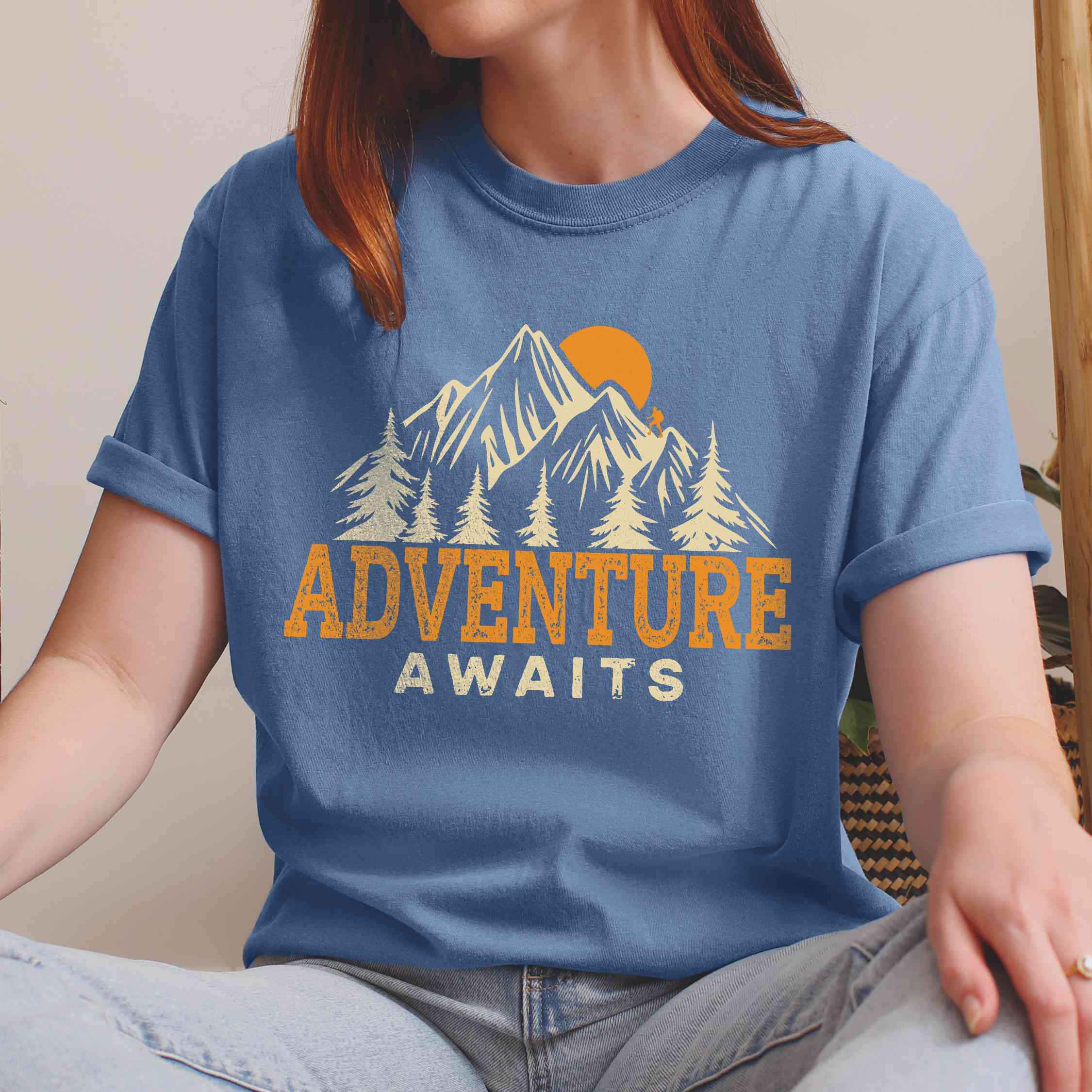 Woman wearing blue jean "Adventure Awaits" colored statement tee with bold typography, mountains, birds, perfect for adventure motivated individuals.