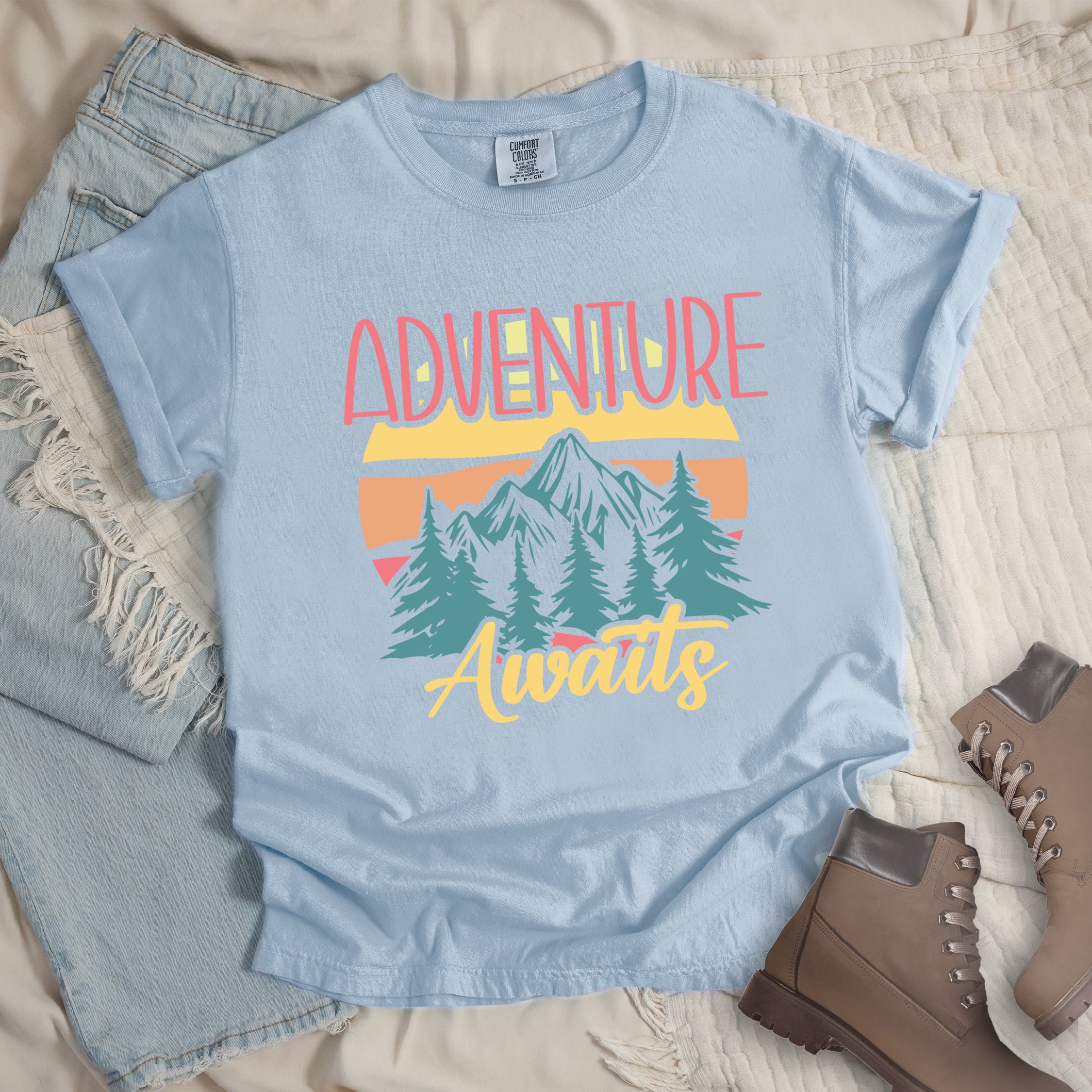 Chambray (Light Blue) t-shirt with retro-style 'Adventure Awaits' design featuring pink gradient text above mountain landscape with teal pine trees, colorful sunset stripes in yellow, orange and pink, and yellow script 'Awaits' text below.