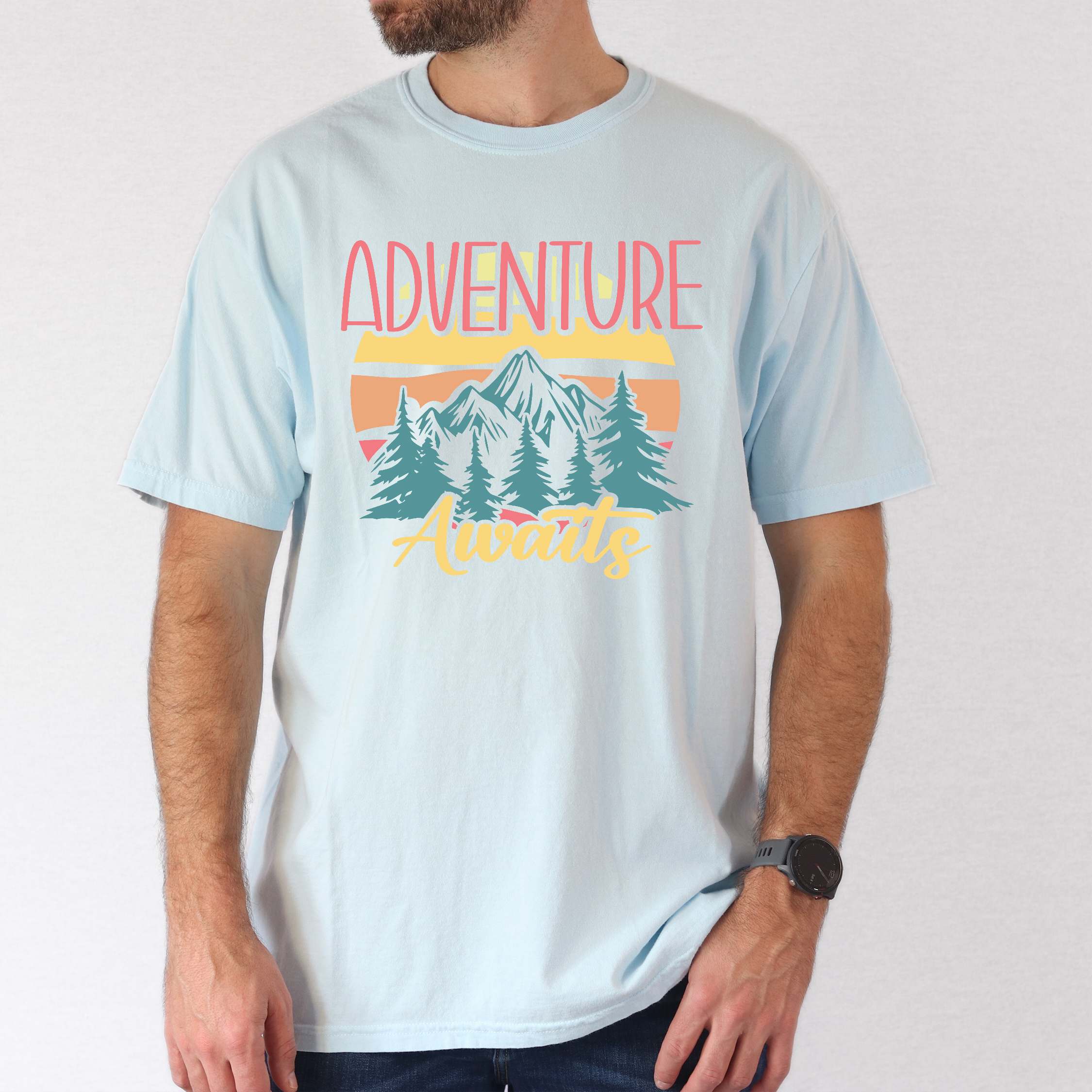 Man wearing chambray "Adventure Awaits" colored statement tee with bold typography, mountains, birds, perfect for adventure motivated individuals.