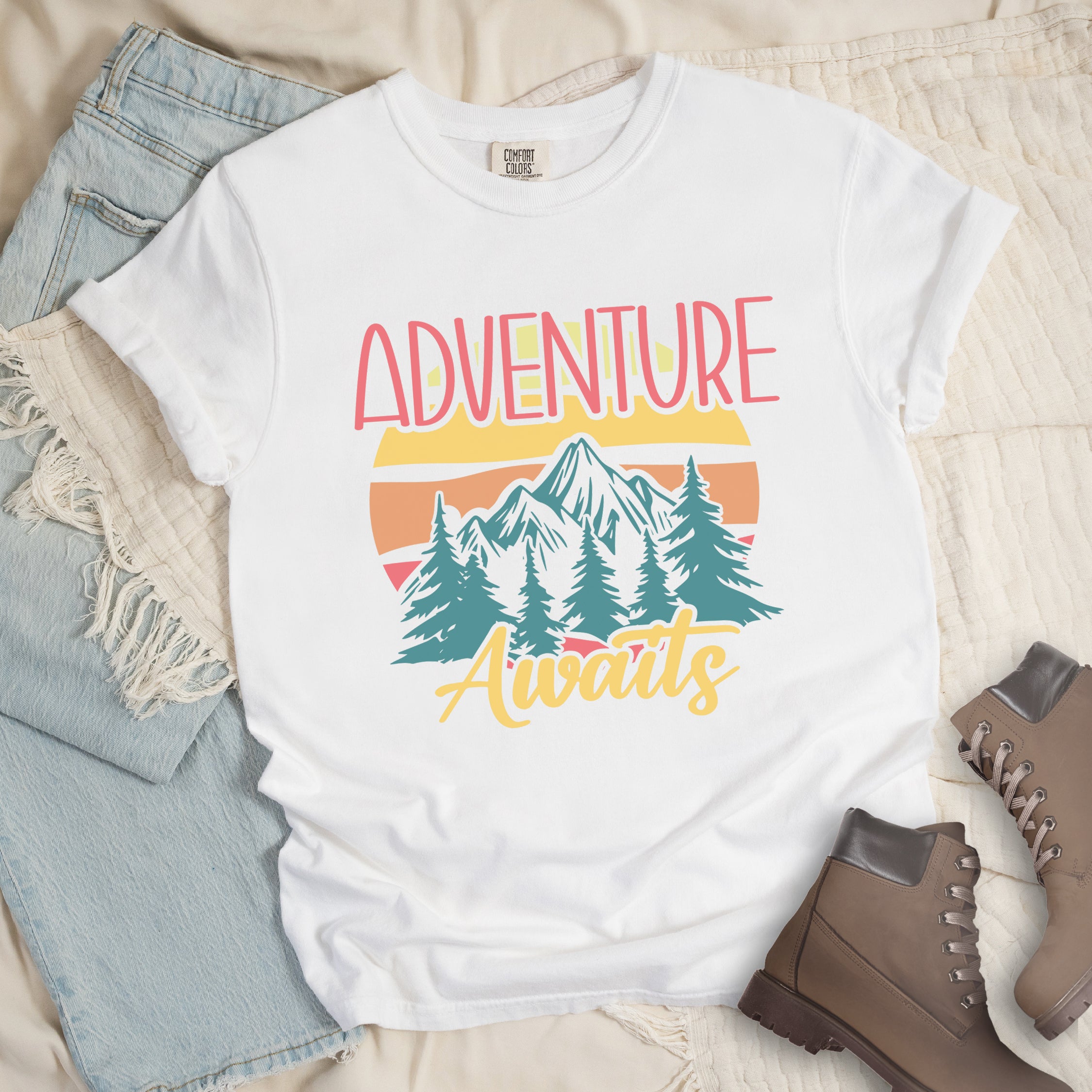 White t-shirt with retro-style 'Adventure Awaits' design featuring pink gradient text above mountain landscape with teal pine trees, colorful sunset stripes in yellow, orange and pink, and yellow script 'Awaits' text below.