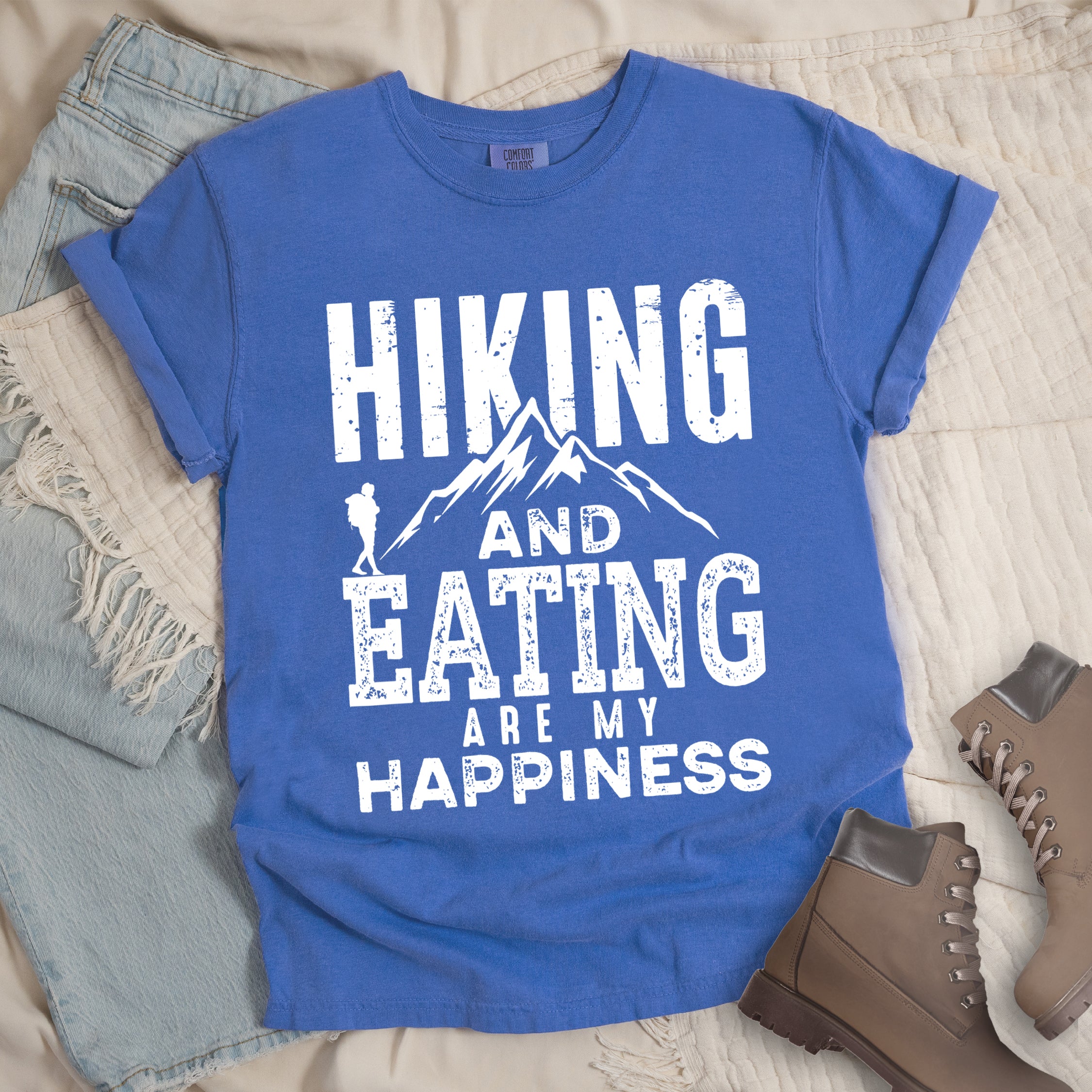Flo Blue colored t-shirt with white distressed text reading 'Hiking and Eating Are My Happiness' featuring mountain silhouette and hiker figure, styled with light wash denim jeans and brown hiking boots on cream bedding.