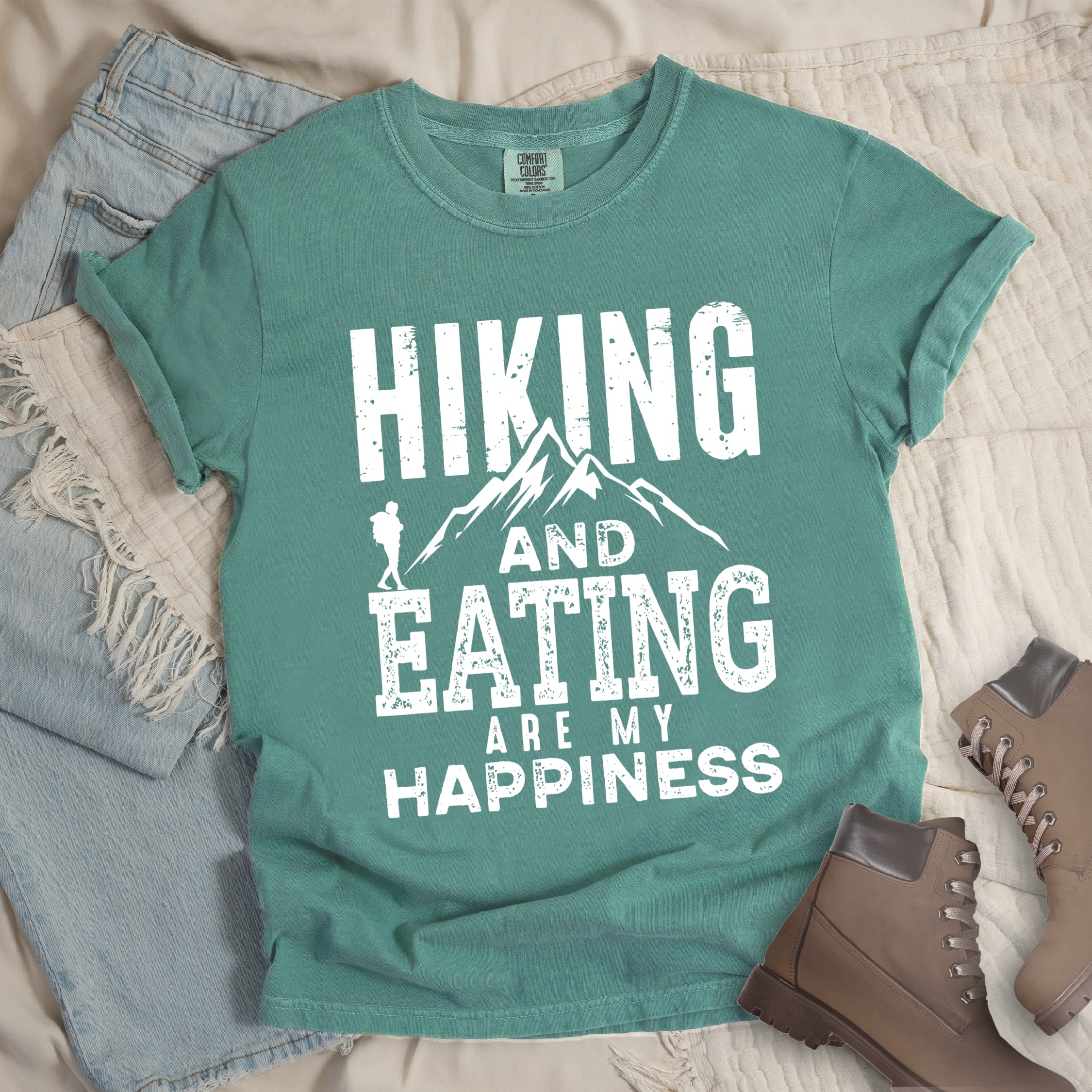 Light Green colored t-shirt with white distressed text reading 'Hiking and Eating Are My Happiness' featuring mountain silhouette and hiker figure, styled with light wash denim jeans and brown hiking boots on cream bedding.