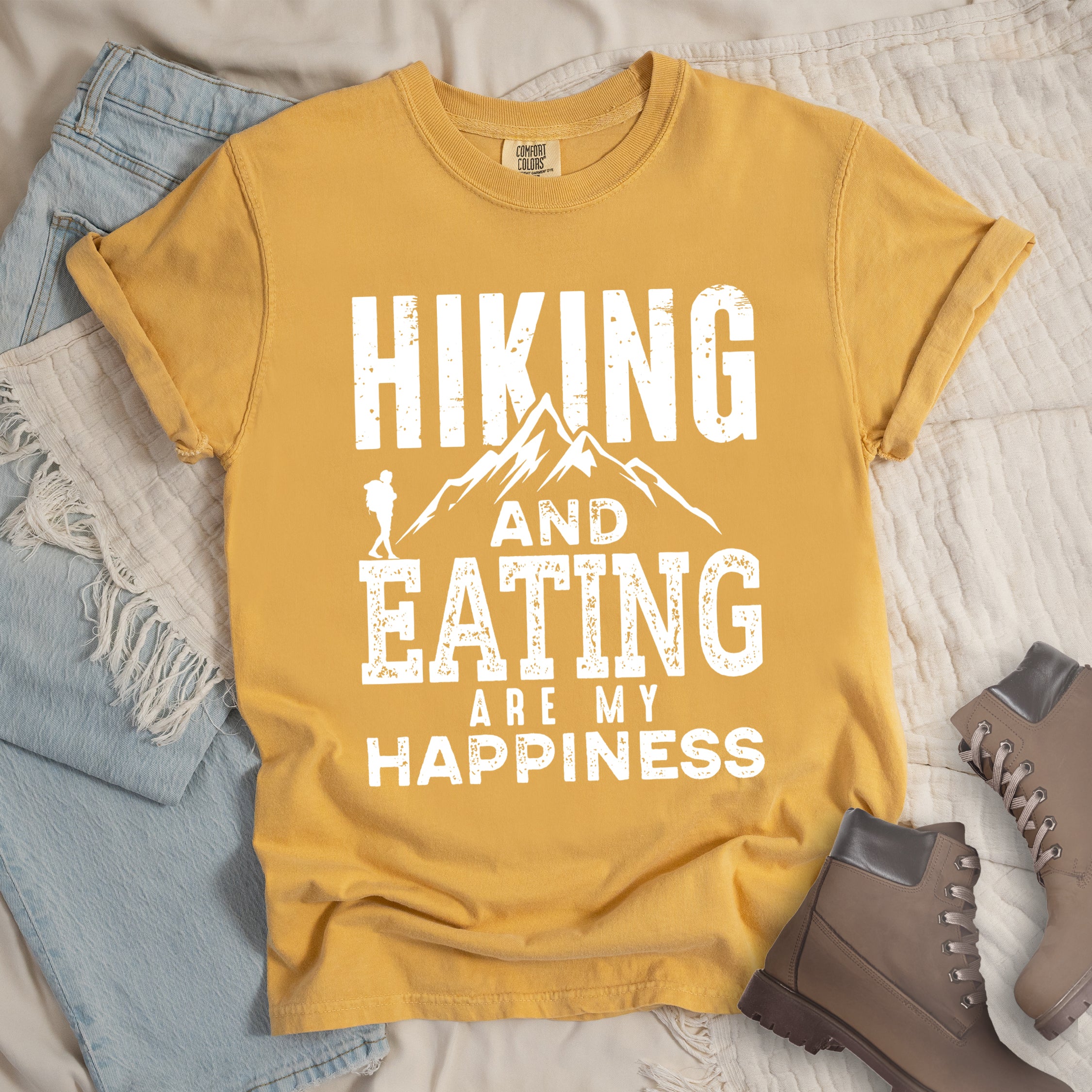 Mustard colored t-shirt with white distressed text reading 'Hiking and Eating Are My Happiness' featuring mountain silhouette and hiker figure, styled with light wash denim jeans and brown hiking boots on cream bedding.