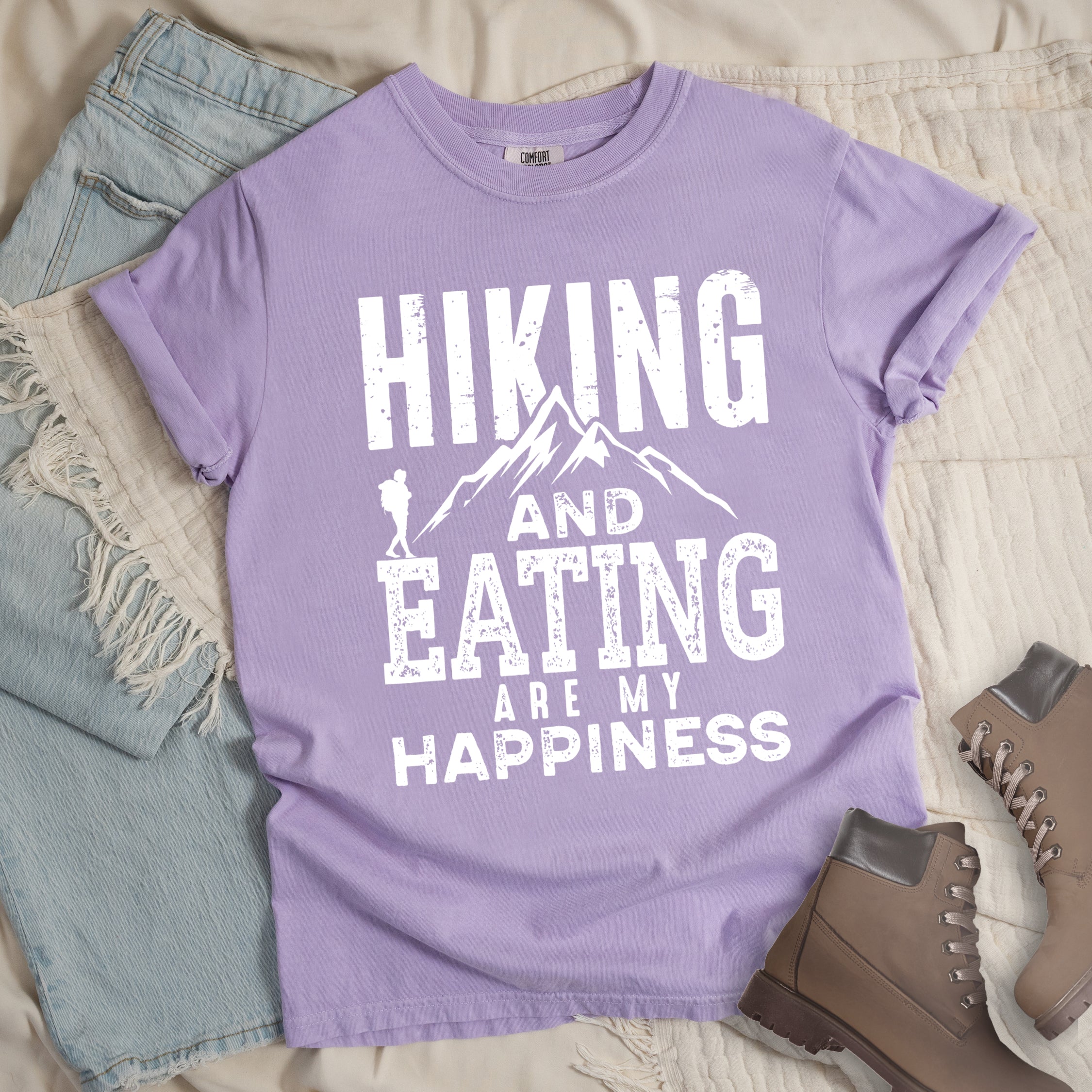 Orchid colored t-shirt with white distressed text reading 'Hiking and Eating Are My Happiness' featuring mountain silhouette and hiker figure, styled with light wash denim jeans and brown hiking boots on cream bedding.