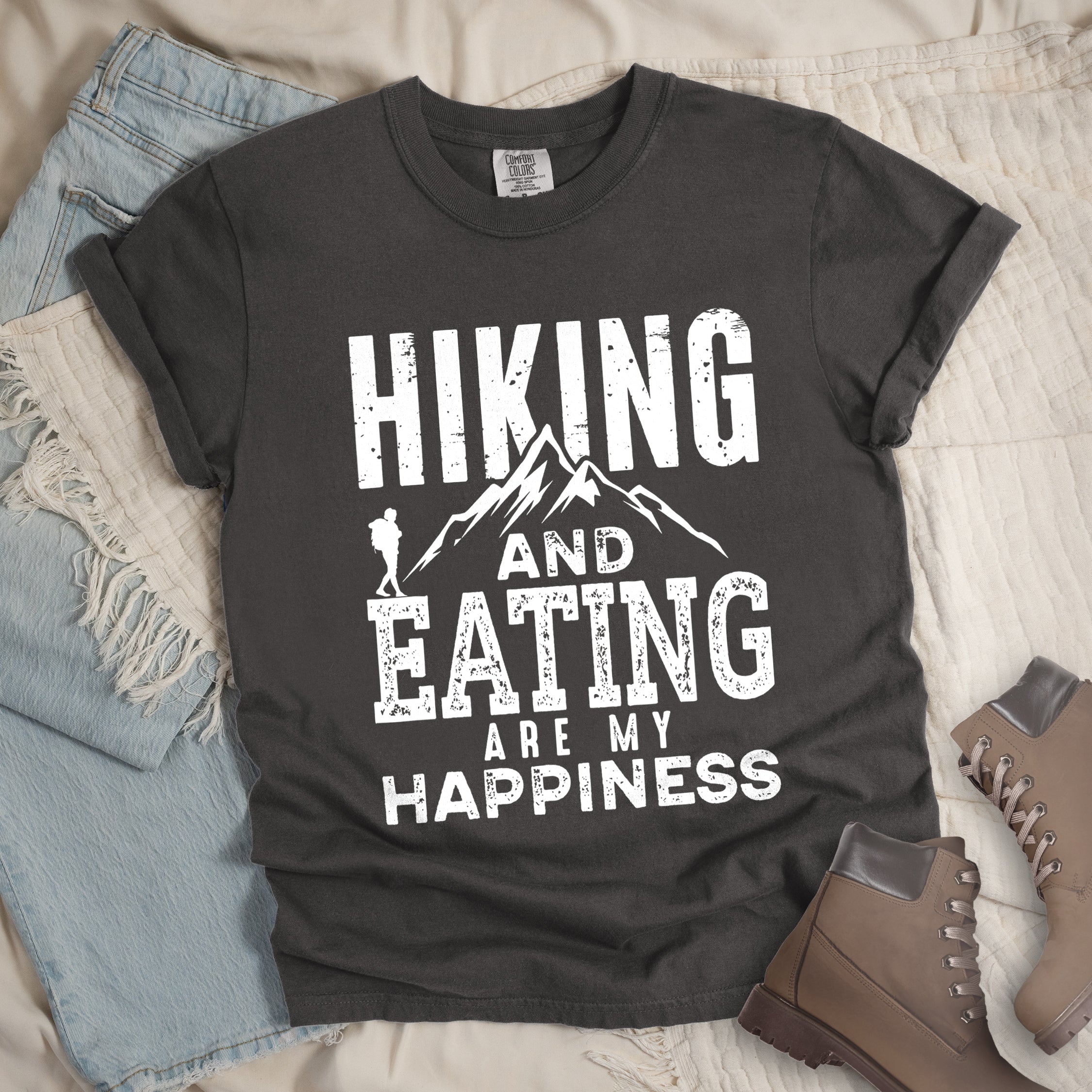 Pepper colored t-shirt with white distressed text reading 'Hiking and Eating Are My Happiness' featuring mountain silhouette and hiker figure, styled with light wash denim jeans and brown hiking boots on cream bedding.