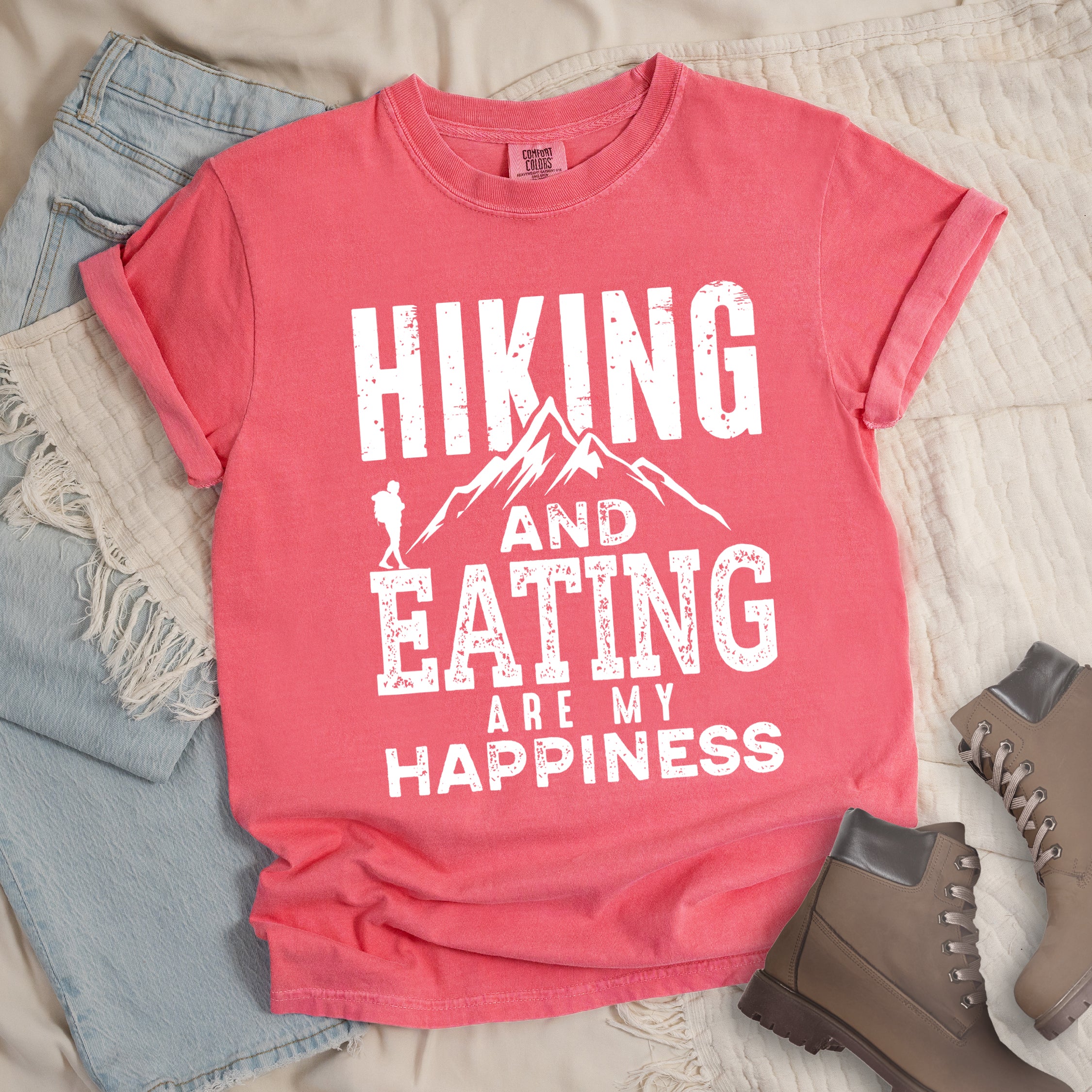 Watermelon colored t-shirt with white distressed text reading 'Hiking and Eating Are My Happiness' featuring mountain silhouette and hiker figure, styled with light wash denim jeans and brown hiking boots on cream bedding.
