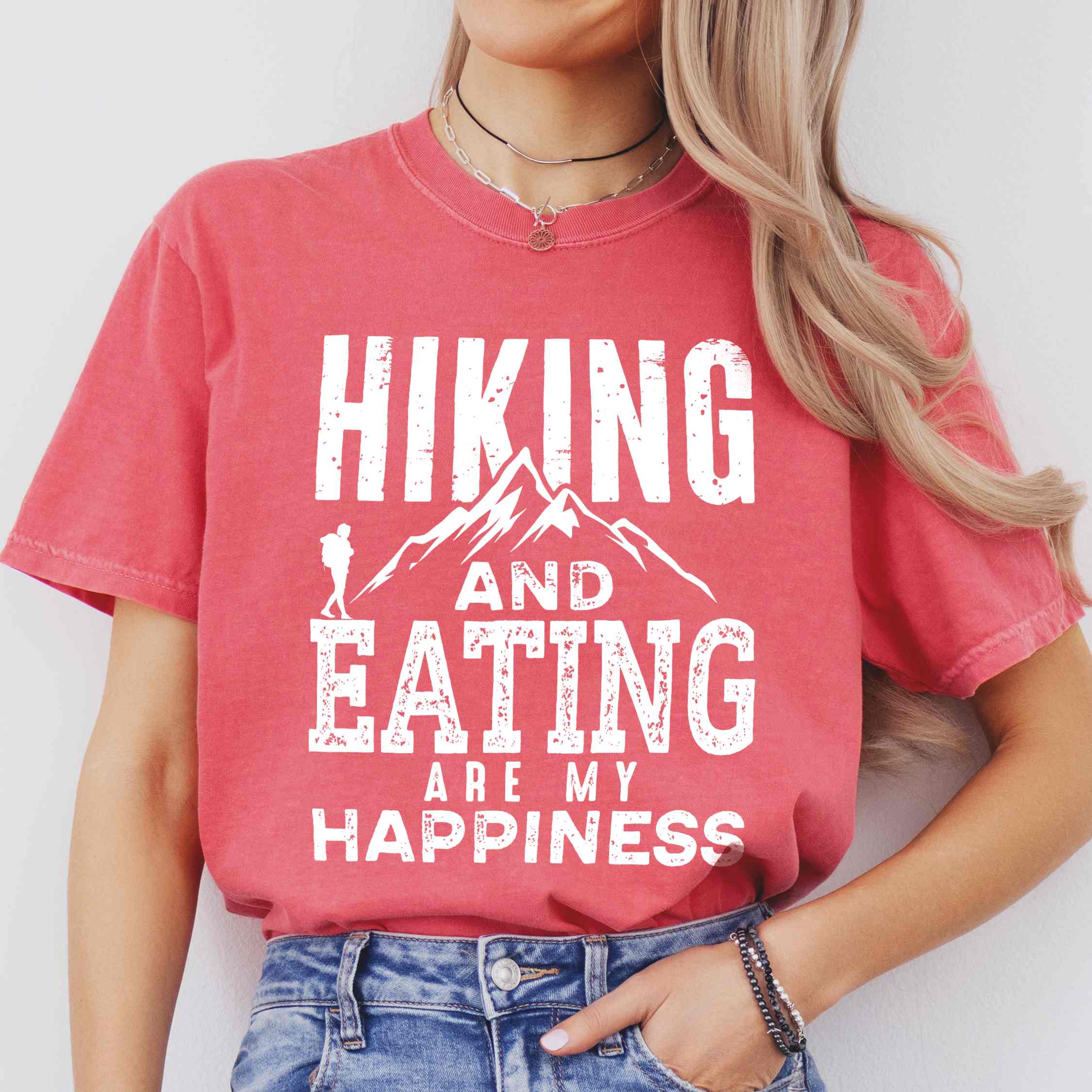 Woman wearing watermelon "Hiking and Eating are my happiness" colored statement tee with bold typography, mountains, birds, perfect for adventure motivated individuals.