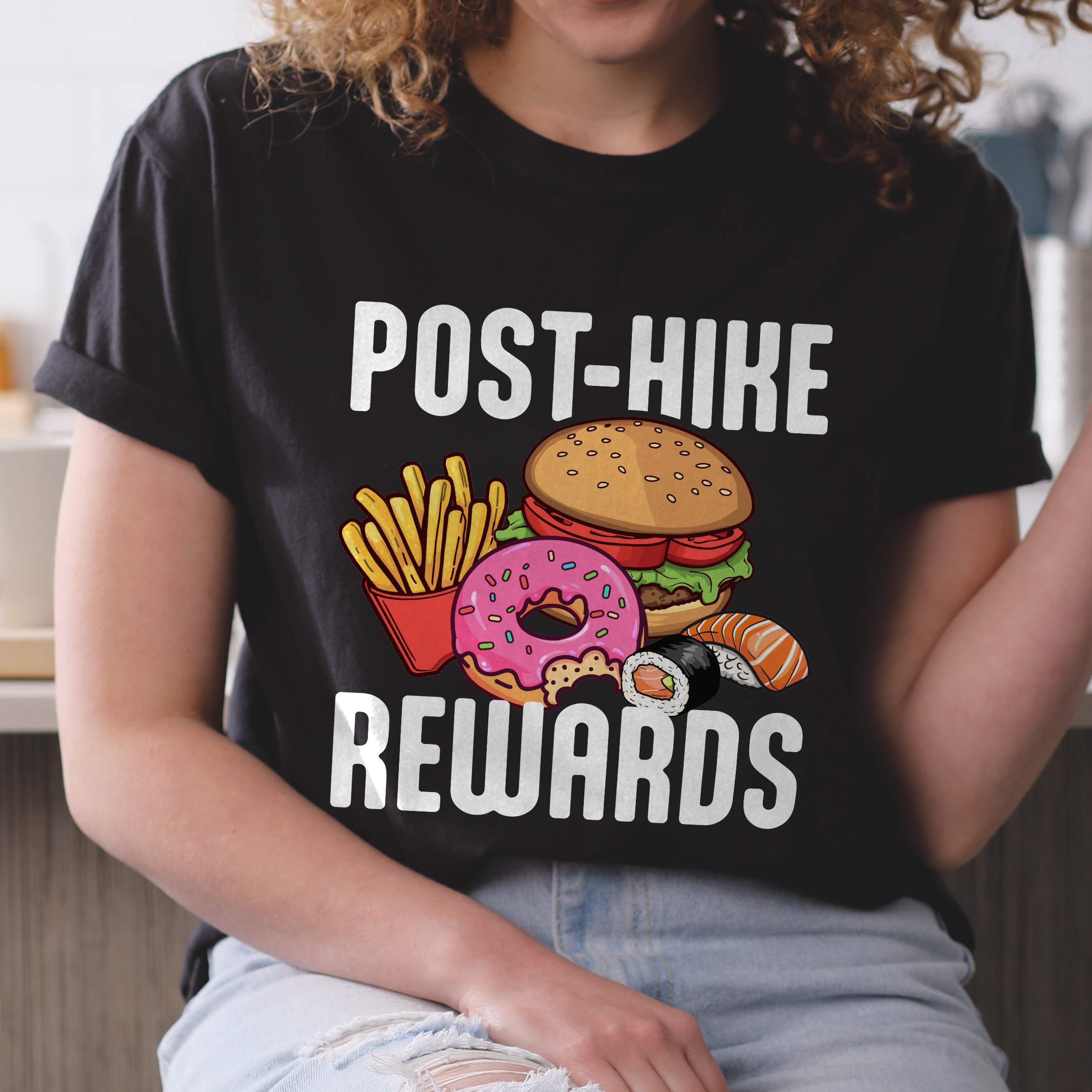 Woman wearing black "Post-hike rewards" colored statement tee with bold typography, mountains, birds, illustrated fast food meal, perfect for adventure motivated individuals and looking for post-hike meal.