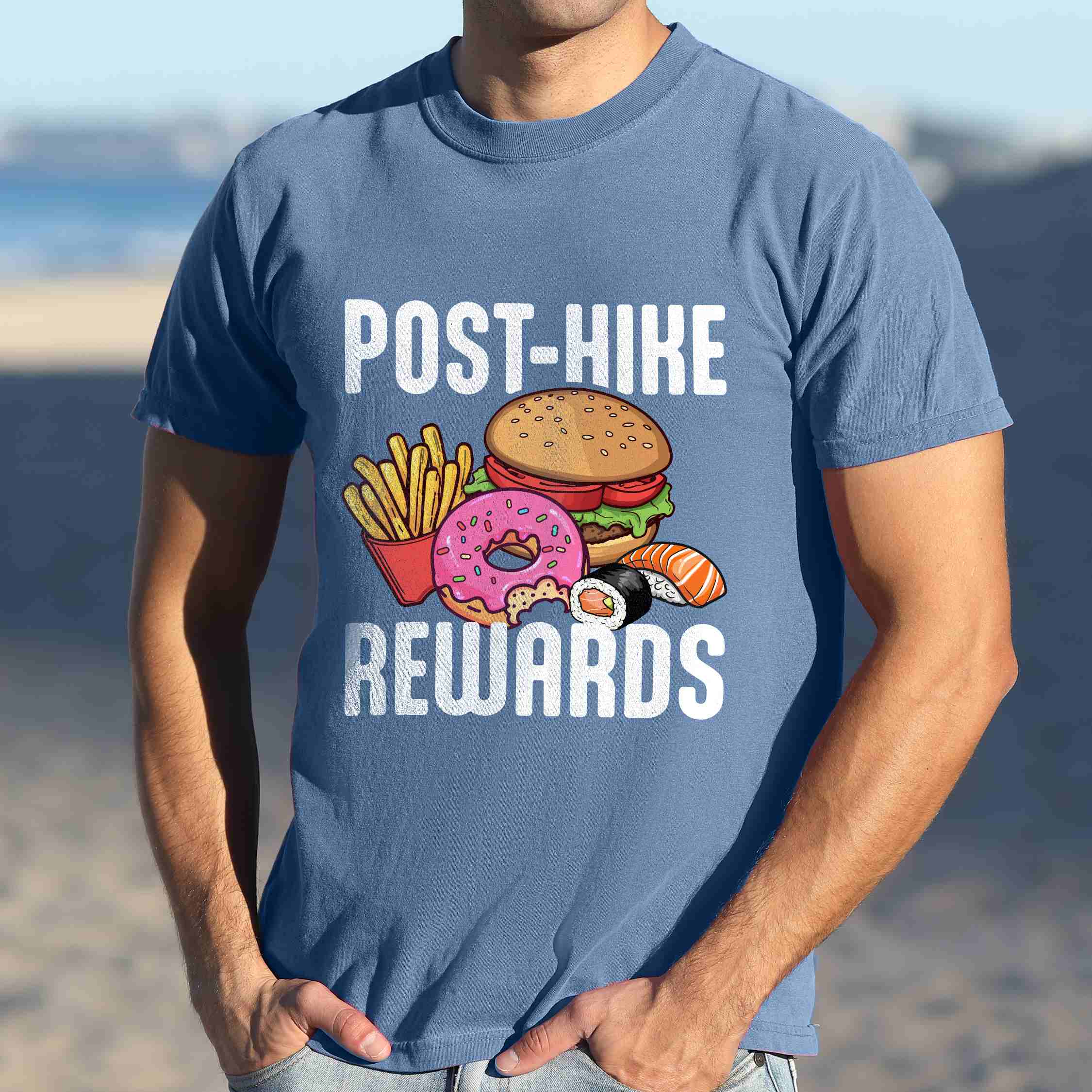 Man wearing blue jean "Post-hike rewards" colored statement tee with bold typography, mountains, birds, illustrated fast food meal, perfect for adventure motivated individuals and looking for post-hike meal.