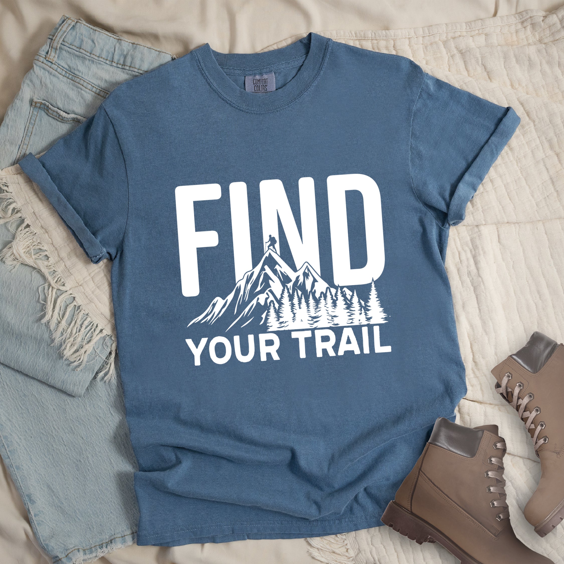 Blue Jean (Denim Blue) graphic t-shirt with 'Find Your Trail' text and white mountain and forest design, styled with light wash jeans and brown hiking boots