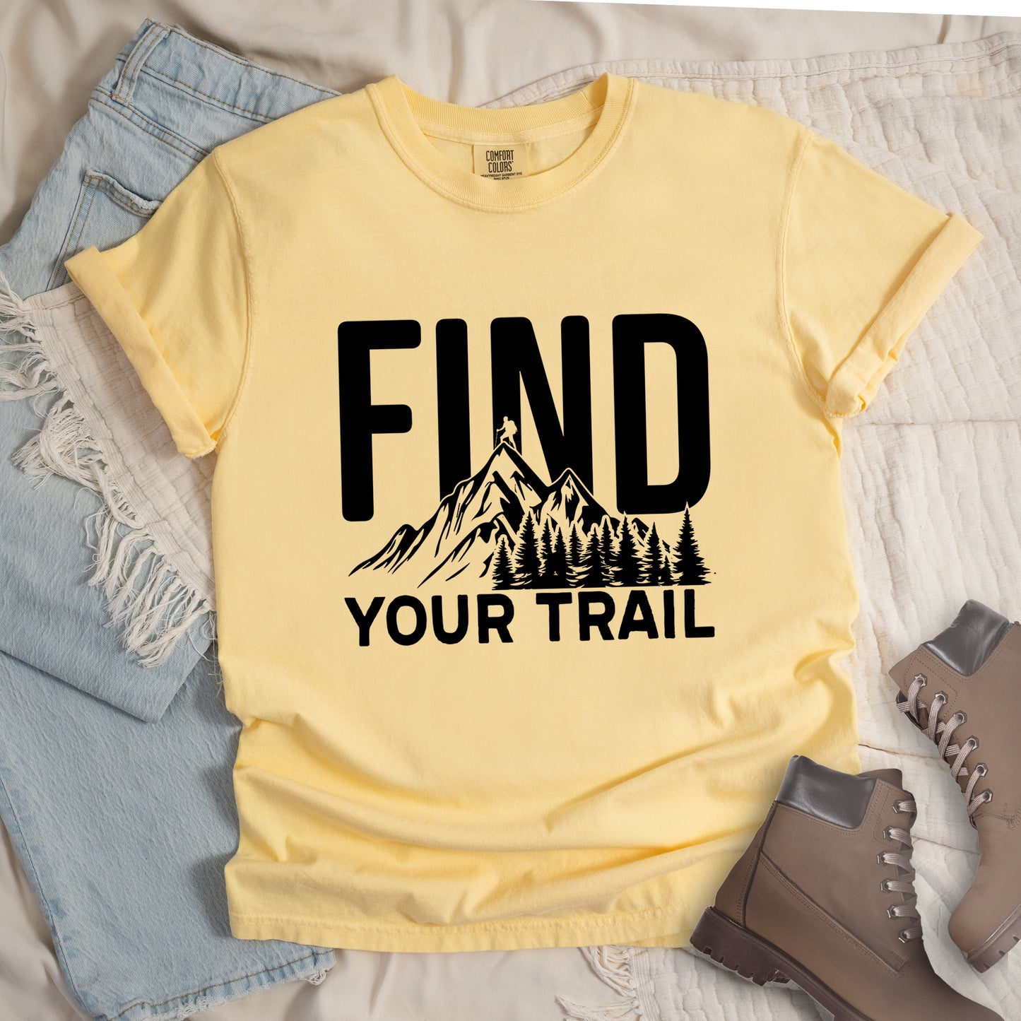 Butter (Light Yellow) graphic t-shirt with 'Find Your Trail' text and black mountain and forest design, styled with light wash jeans and brown hiking boots