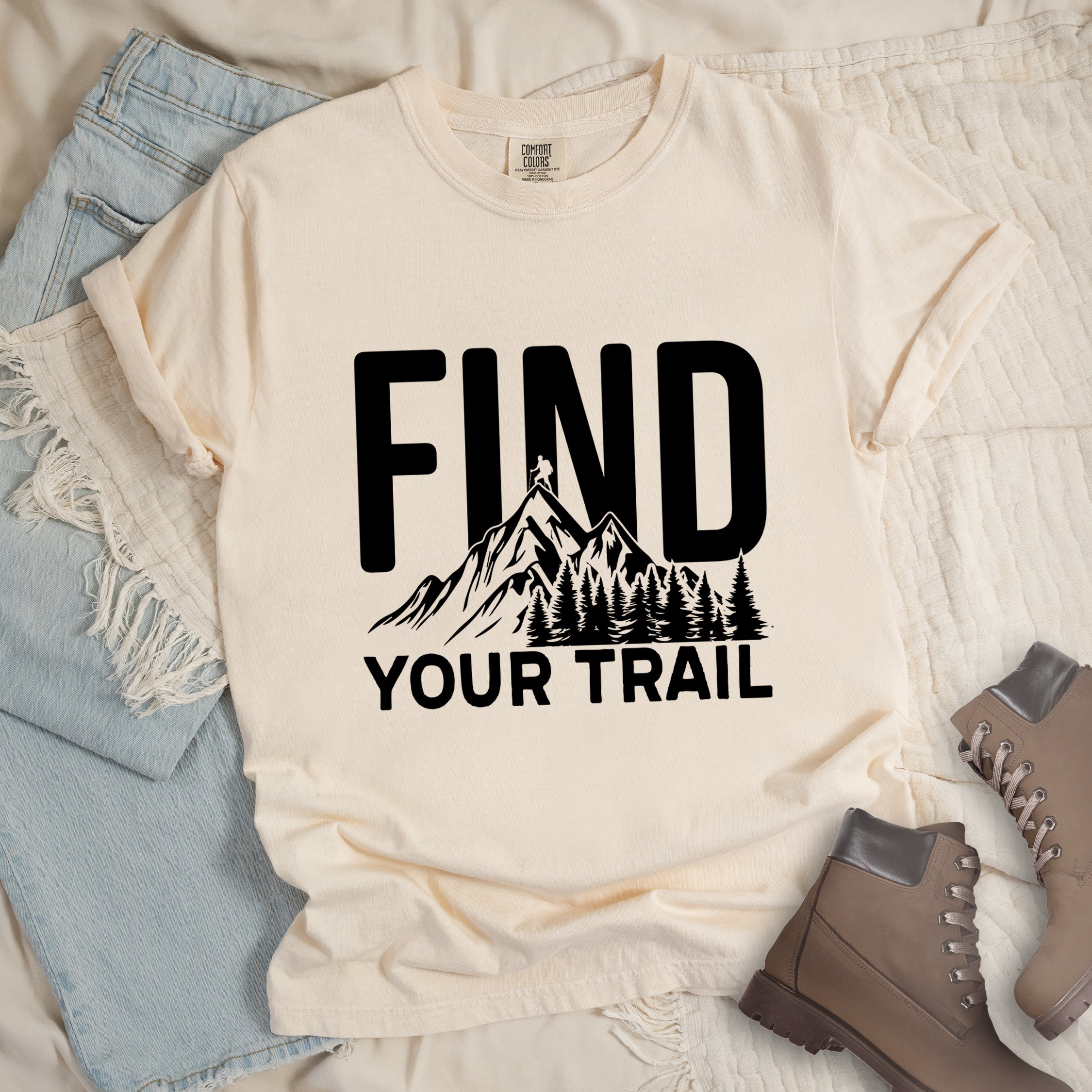 Ivory (Cream White) graphic t-shirt with 'Find Your Trail' text and black mountain and forest design, styled with light wash jeans and brown hiking boots