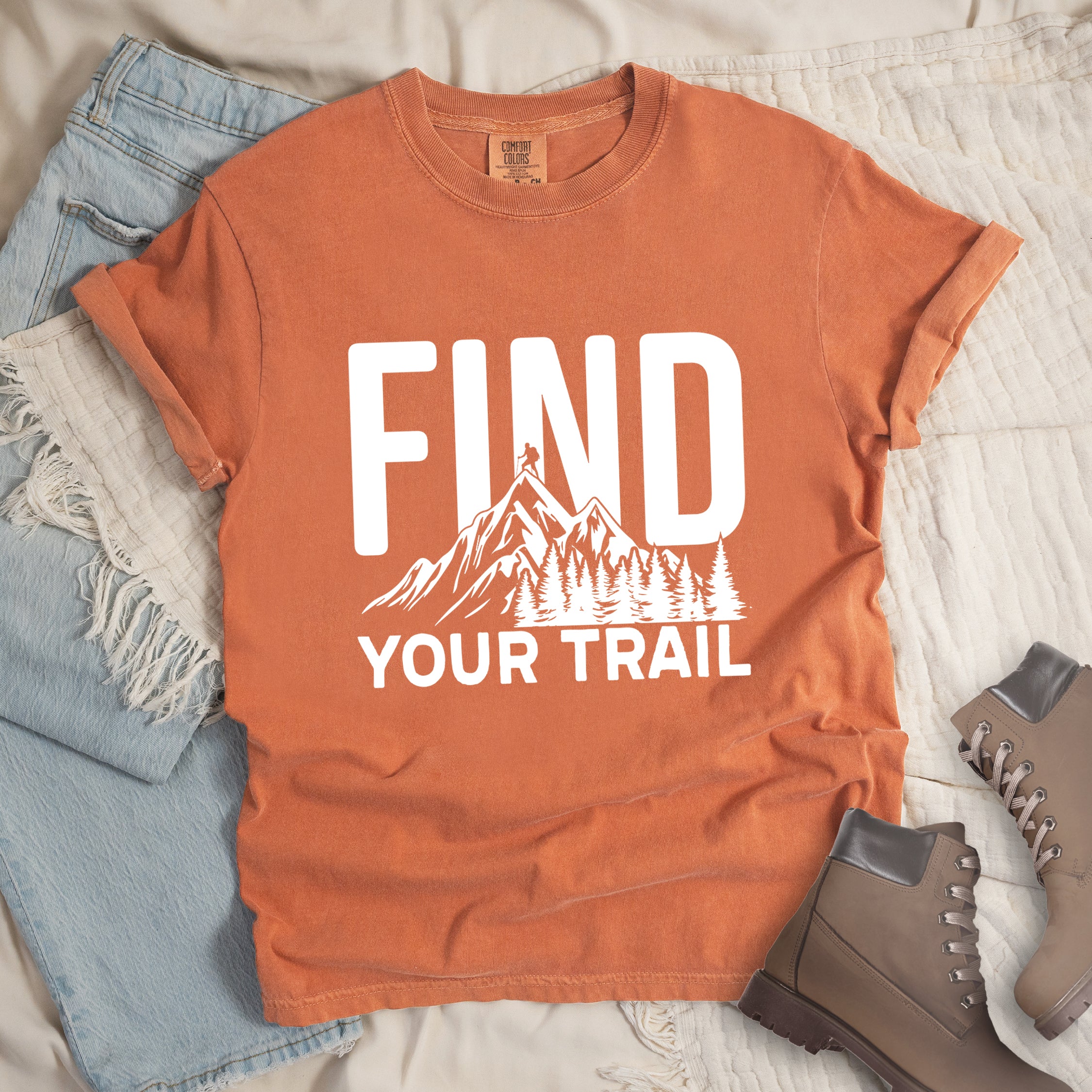 Yam (Brown) graphic t-shirt with 'Find Your Trail' text and white mountain and forest design, styled with light wash jeans and brown hiking boots