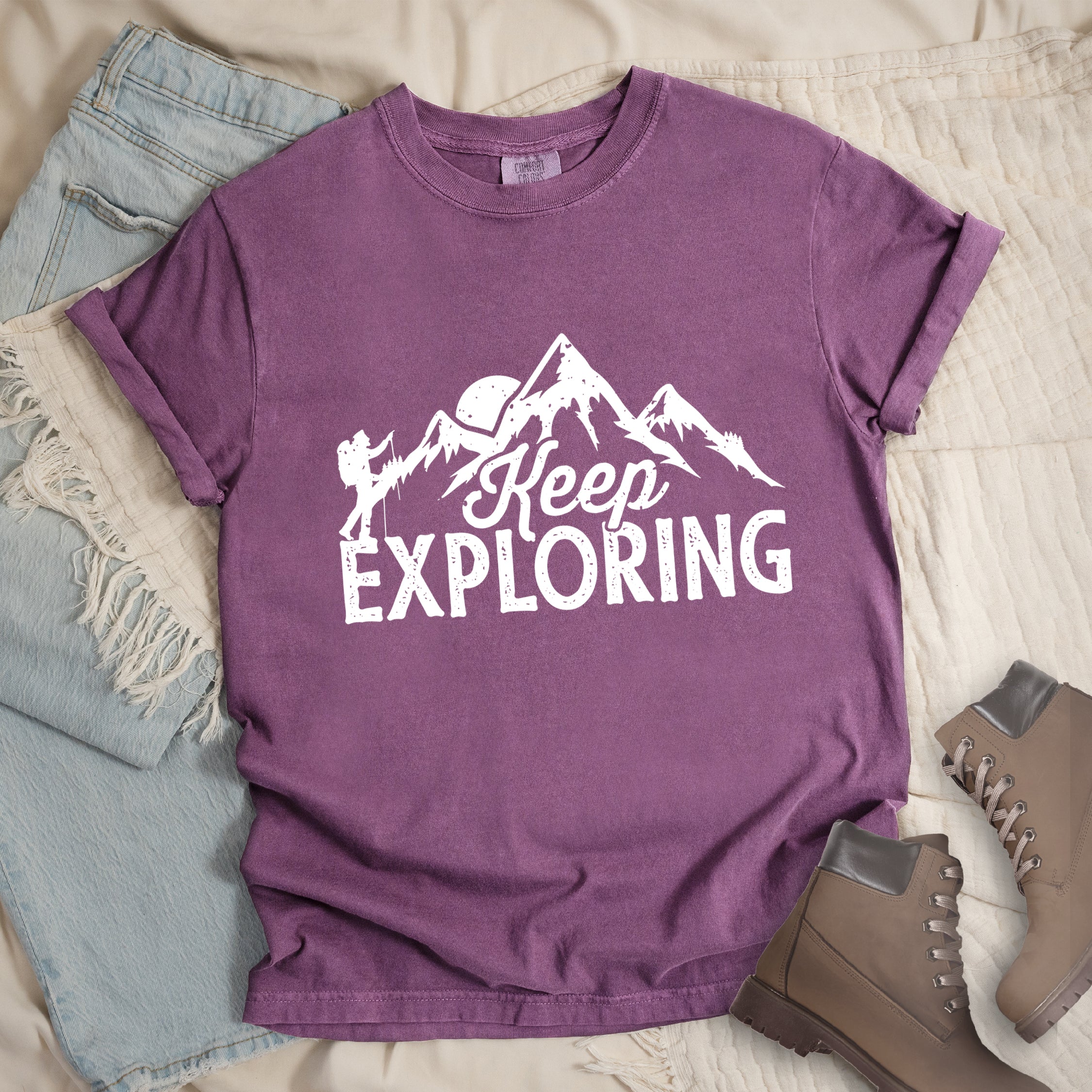 Berry (Purple) colored t-shirt with white distressed 'Keep Exploring' text and mountain range silhouette featuring hiker figure and moon, styled with light wash denim jeans and brown hiking boots on cream bedding.
