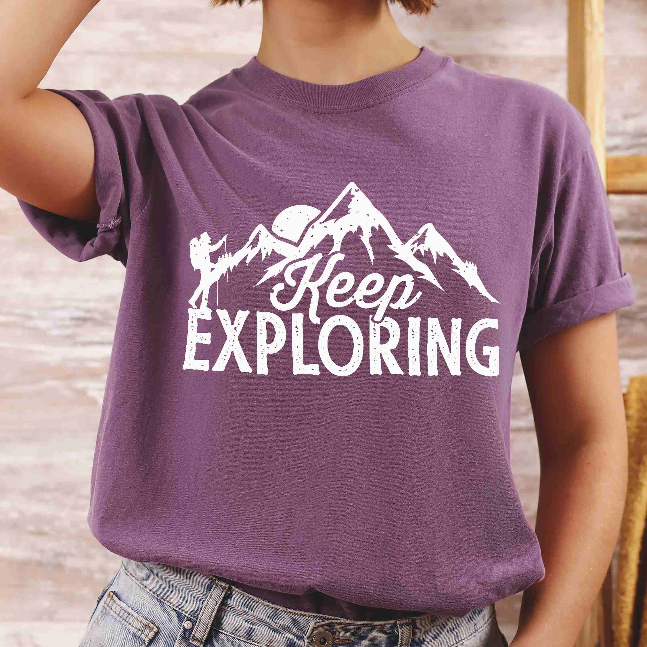 Woman wearing berry "Keep Exploring" colored statement tee with bold typography, mountains, birds, perfect for adventure motivated individuals.