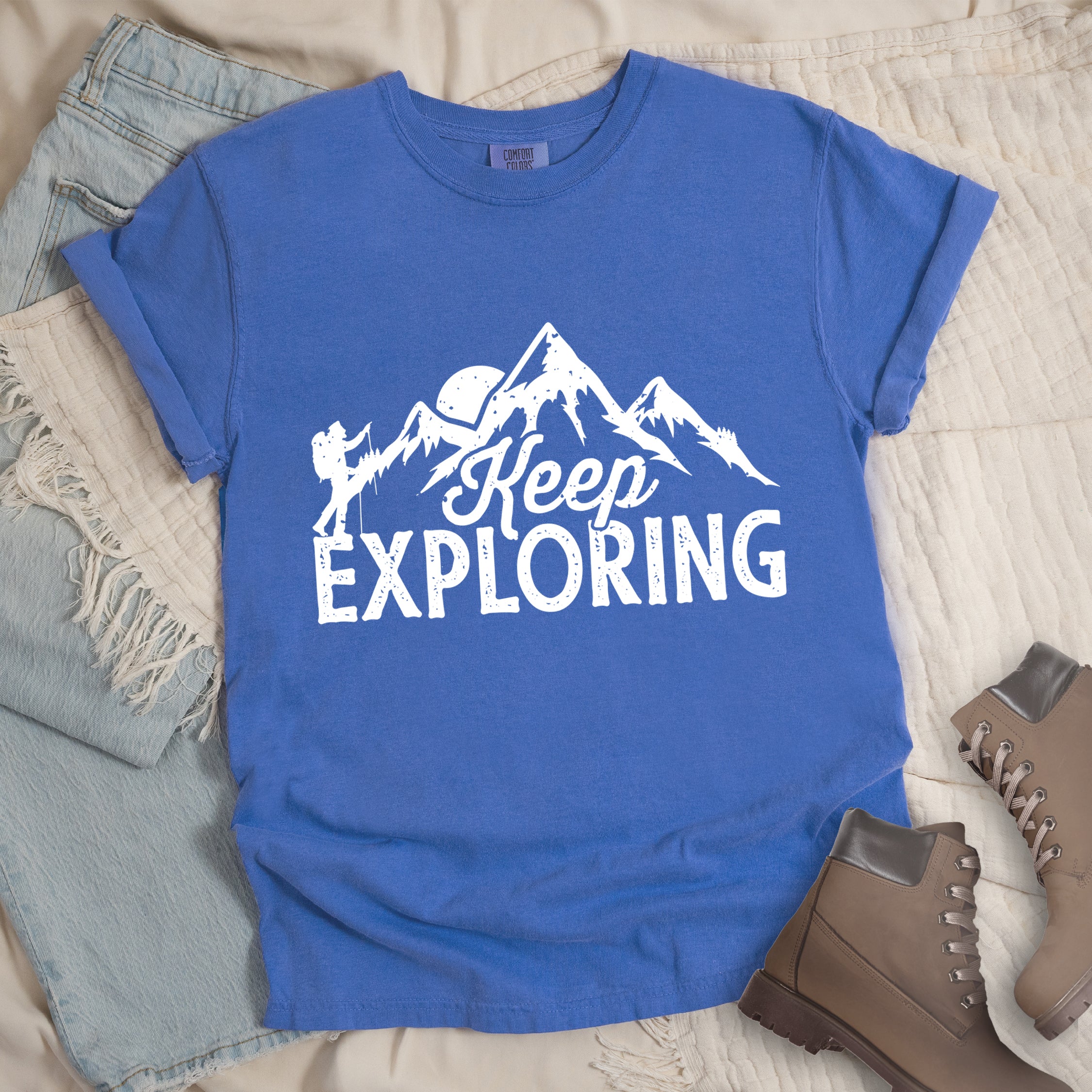 Flo Blue colored t-shirt with white distressed 'Keep Exploring' text and mountain range silhouette featuring hiker figure and moon, styled with light wash denim jeans and brown hiking boots on cream bedding.