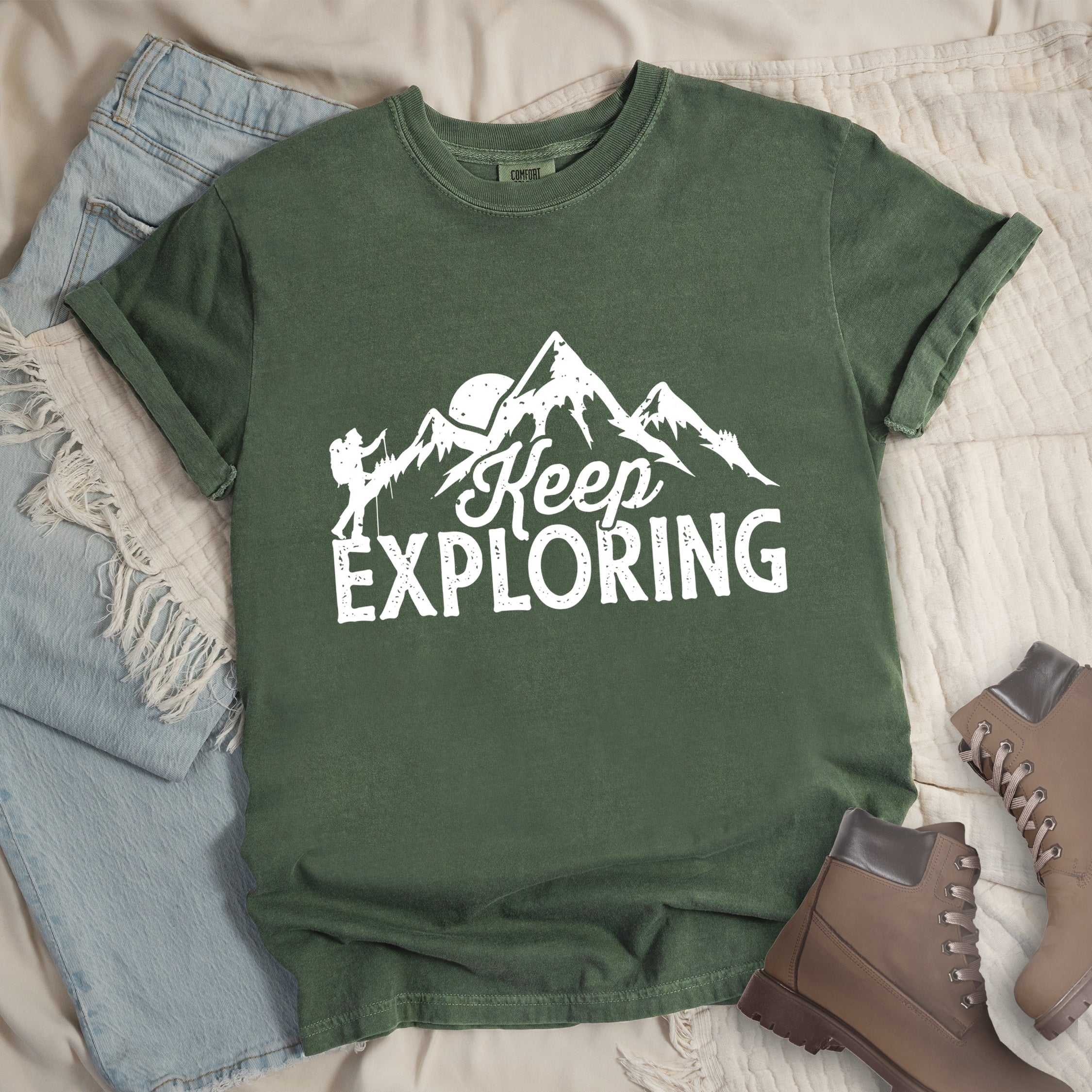 Moss colored (Forest green) t-shirt with white distressed 'Keep Exploring' text and mountain range silhouette featuring hiker figure and moon, styled with light wash denim jeans and brown hiking boots on cream bedding.