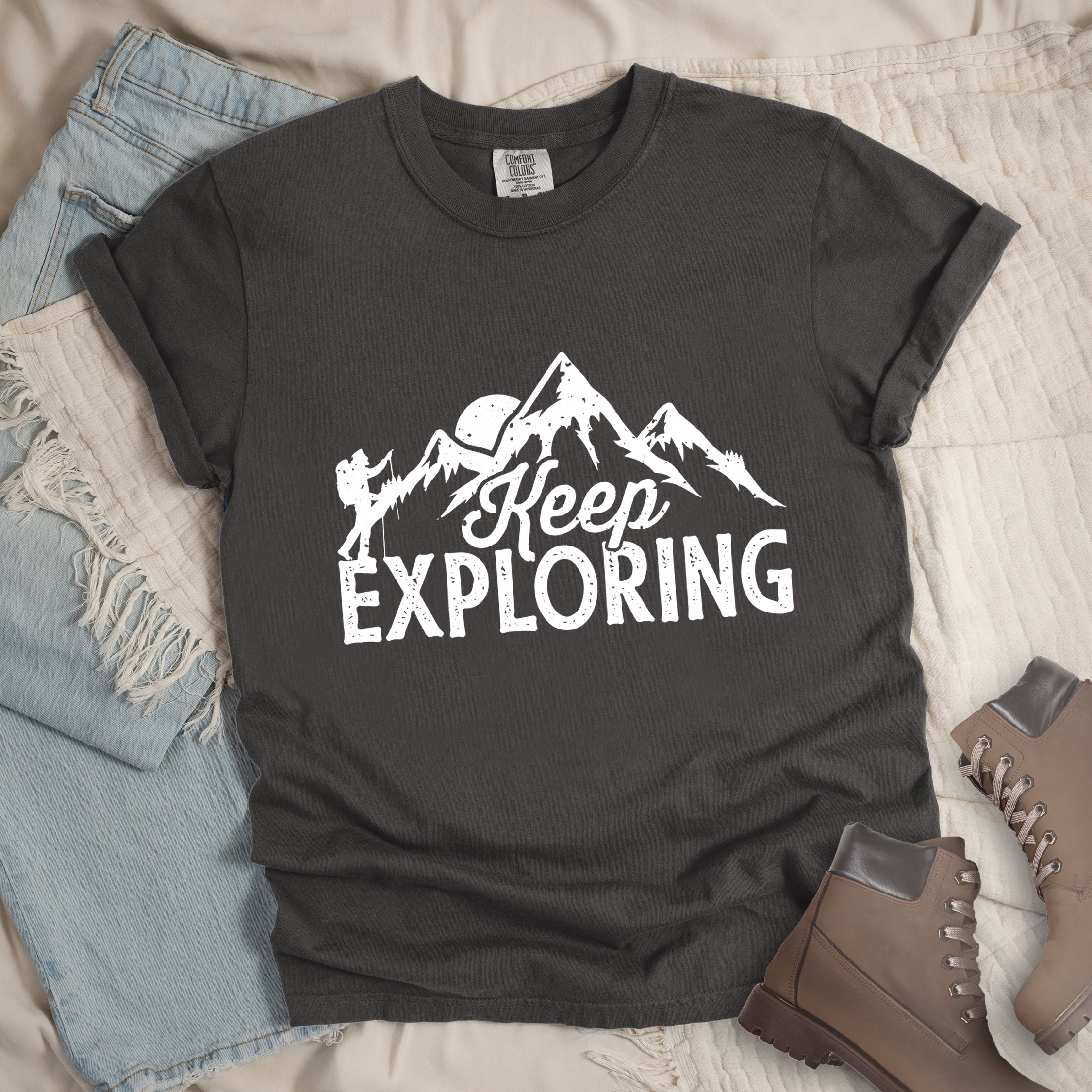 Pepper colored t-shirt with white distressed 'Keep Exploring' text and mountain range silhouette featuring hiker figure and moon, styled with light wash denim jeans and brown hiking boots on cream bedding.