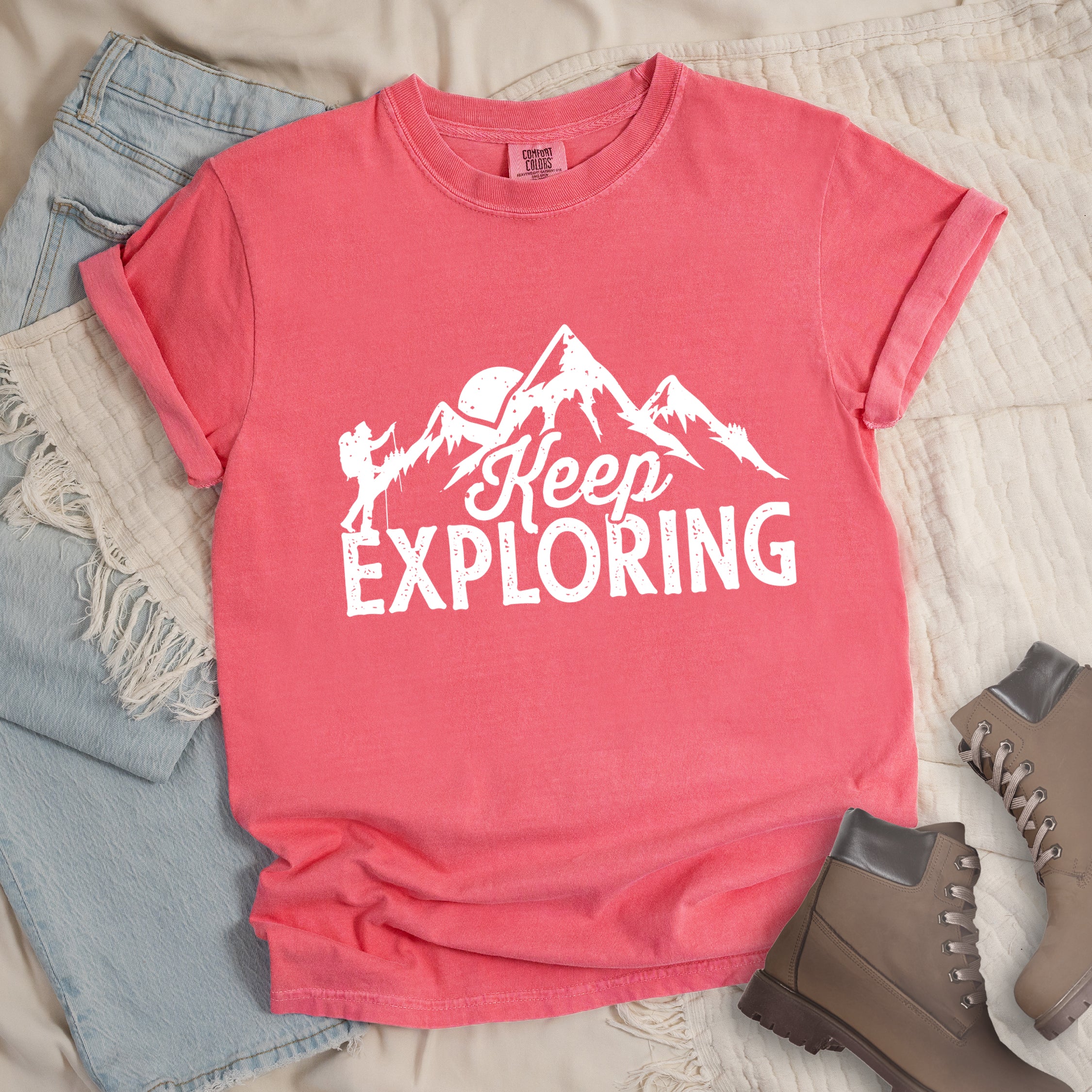 Watermelon colored t-shirt with white distressed 'Keep Exploring' text and mountain range silhouette featuring hiker figure and moon, styled with light wash denim jeans and brown hiking boots on cream bedding.