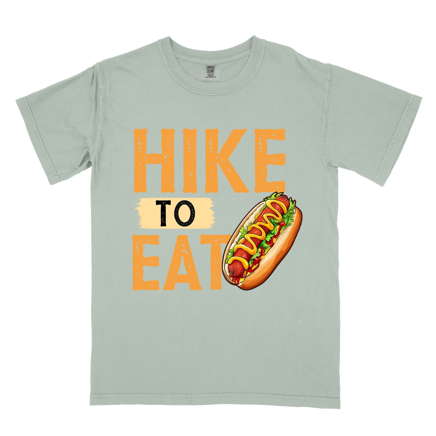 Bay (Light Gray/Green) t-shirt with "HIKE TO EAT" in distressed orange lettering. The word "TO" appears on a beige background between "HIKE" and "EAT". A colorful hot dog illustration with mustard, ketchup and toppings appears next to the "T" in "EAT".
