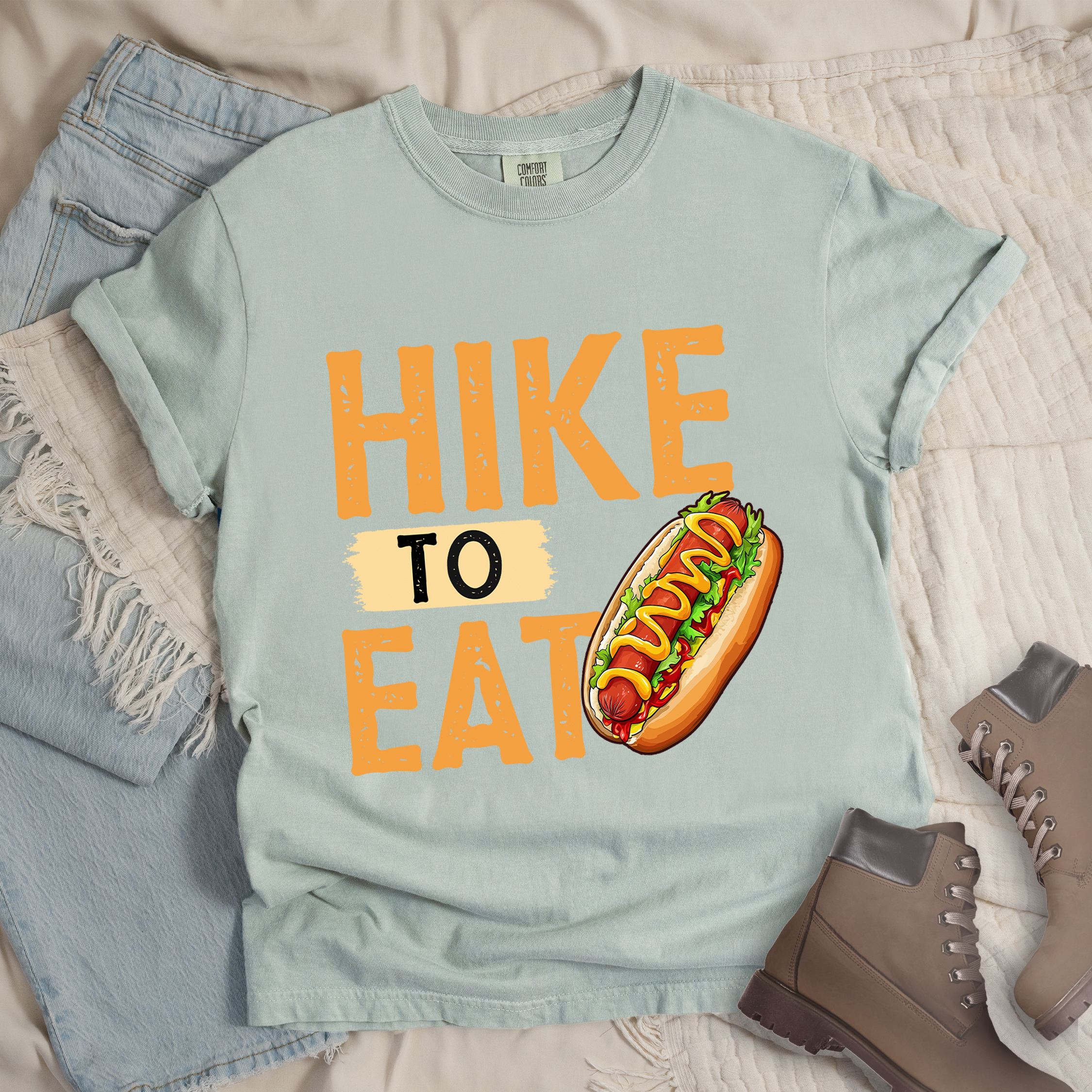 Light grey shirt showing a hiking design with 'HIKE TO EAT phrase and an illustration of a hotdog in a bun with mustard on top