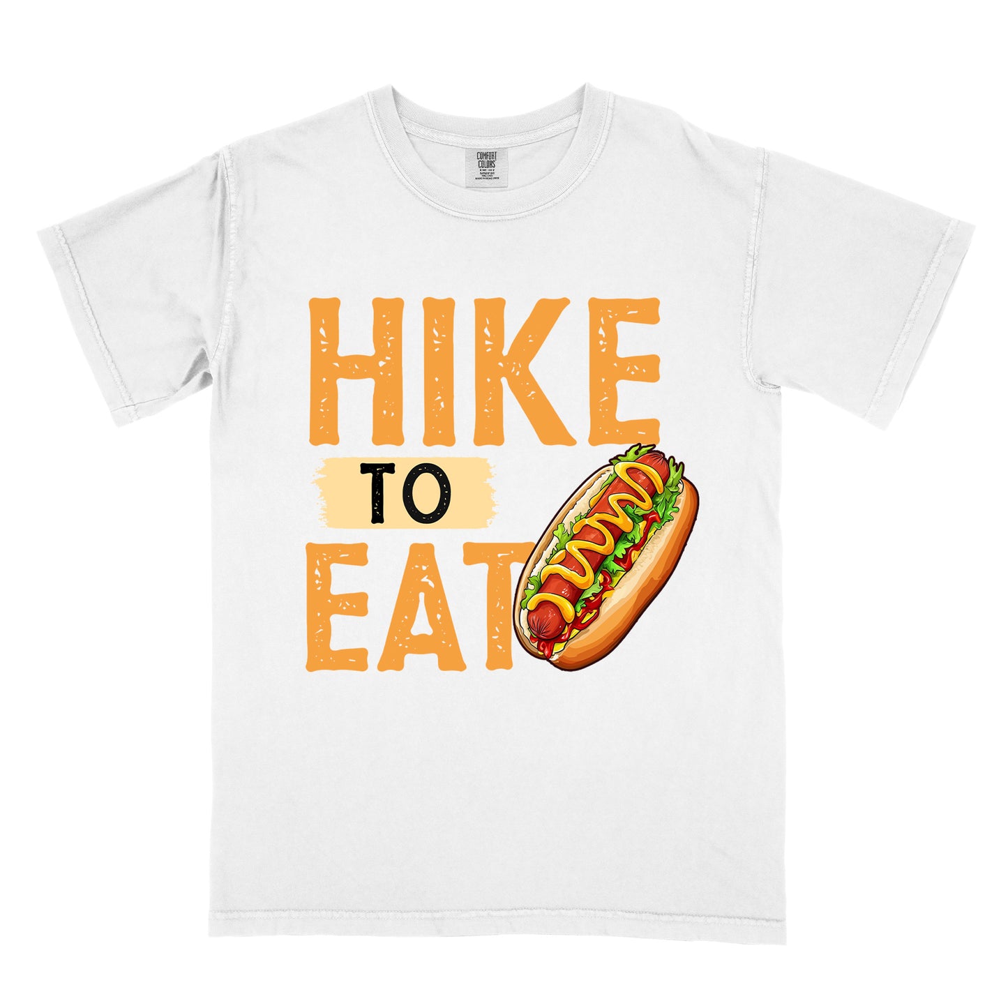 White t-shirt with "HIKE TO EAT" in distressed orange lettering. The word "TO" appears on a beige background between "HIKE" and "EAT". A colorful hot dog illustration with mustard, ketchup and toppings appears next to the "T" in "EAT".