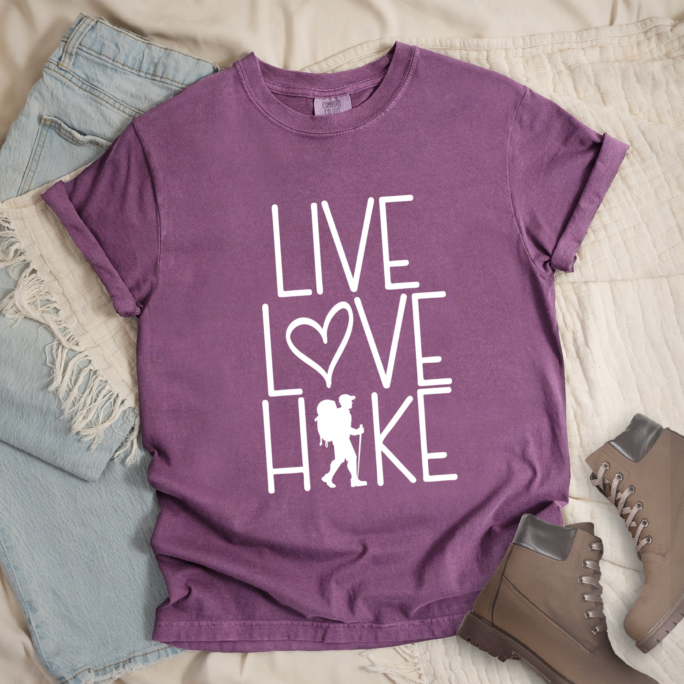 Berry (violet) colored t-shirt with white text reading 'Live Love Hike' featuring a heart symbol and a male hiker silhouette with backpack, styled with light wash denim jeans and brown hiking boots on white bedding.