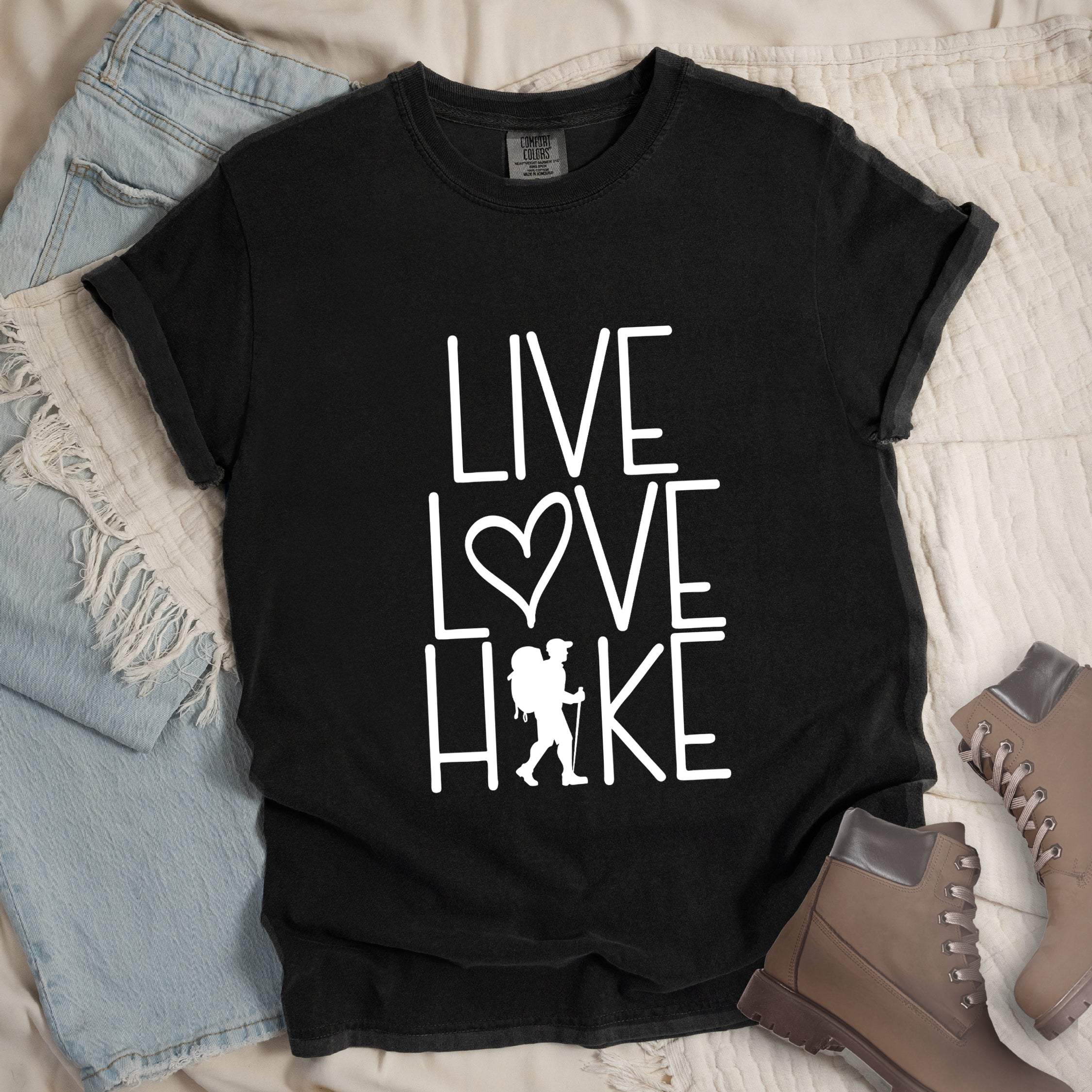 Black colored t-shirt with white text reading 'Live Love Hike' featuring a heart symbol and a male hiker silhouette with backpack, styled with light wash denim jeans and brown hiking boots on white bedding.