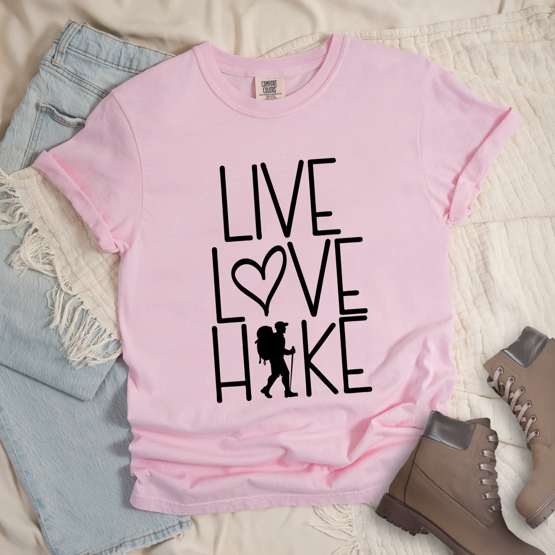 Blossom (pink) colored t-shirt with black text reading 'Live Love Hike' featuring a heart symbol and a male hiker silhouette with backpack, styled with light wash denim jeans and brown hiking boots on white bedding.