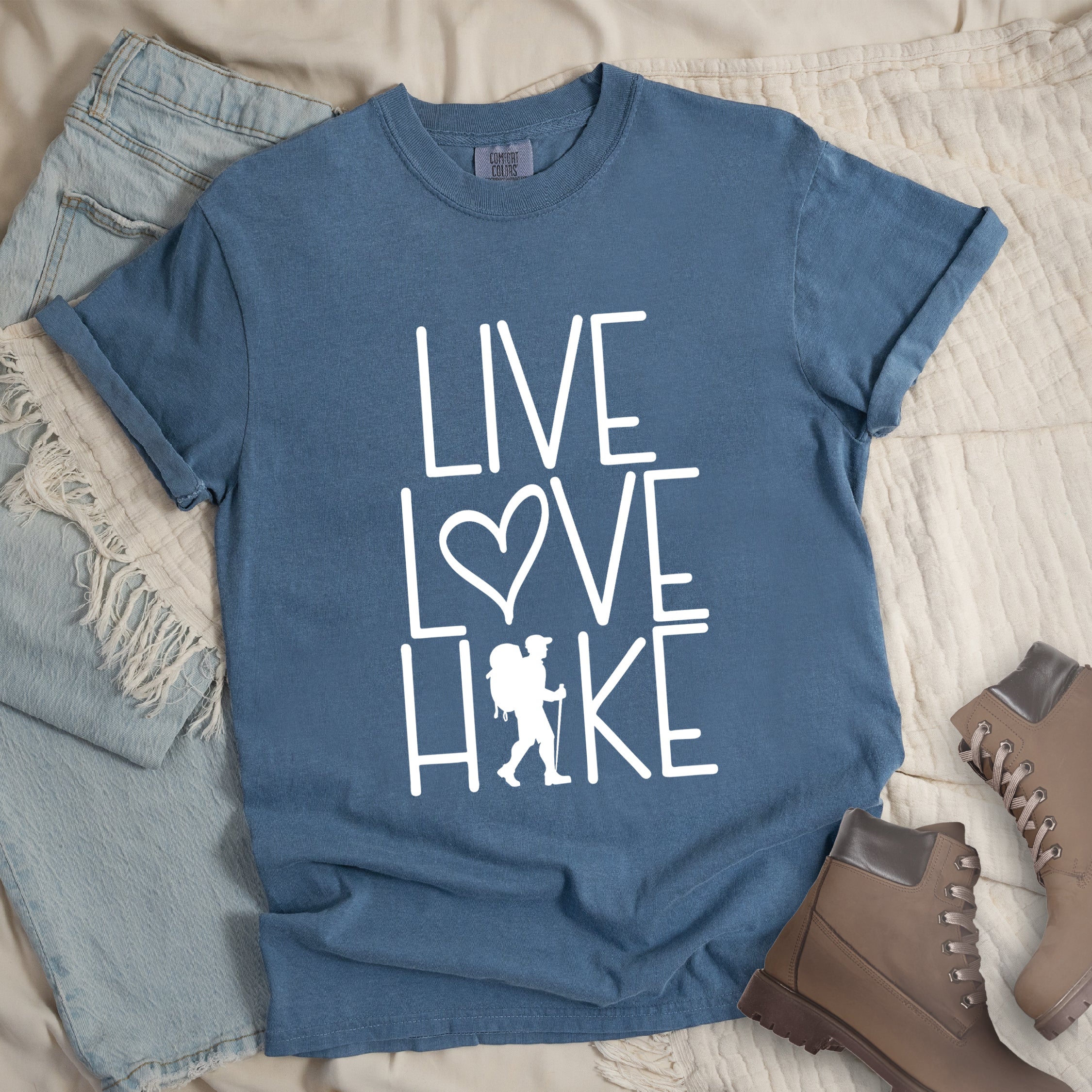 Blue Jean colored t-shirt with white text reading 'Live Love Hike' featuring a heart symbol and a male hiker silhouette with backpack, styled with light wash denim jeans and brown hiking boots on white bedding.