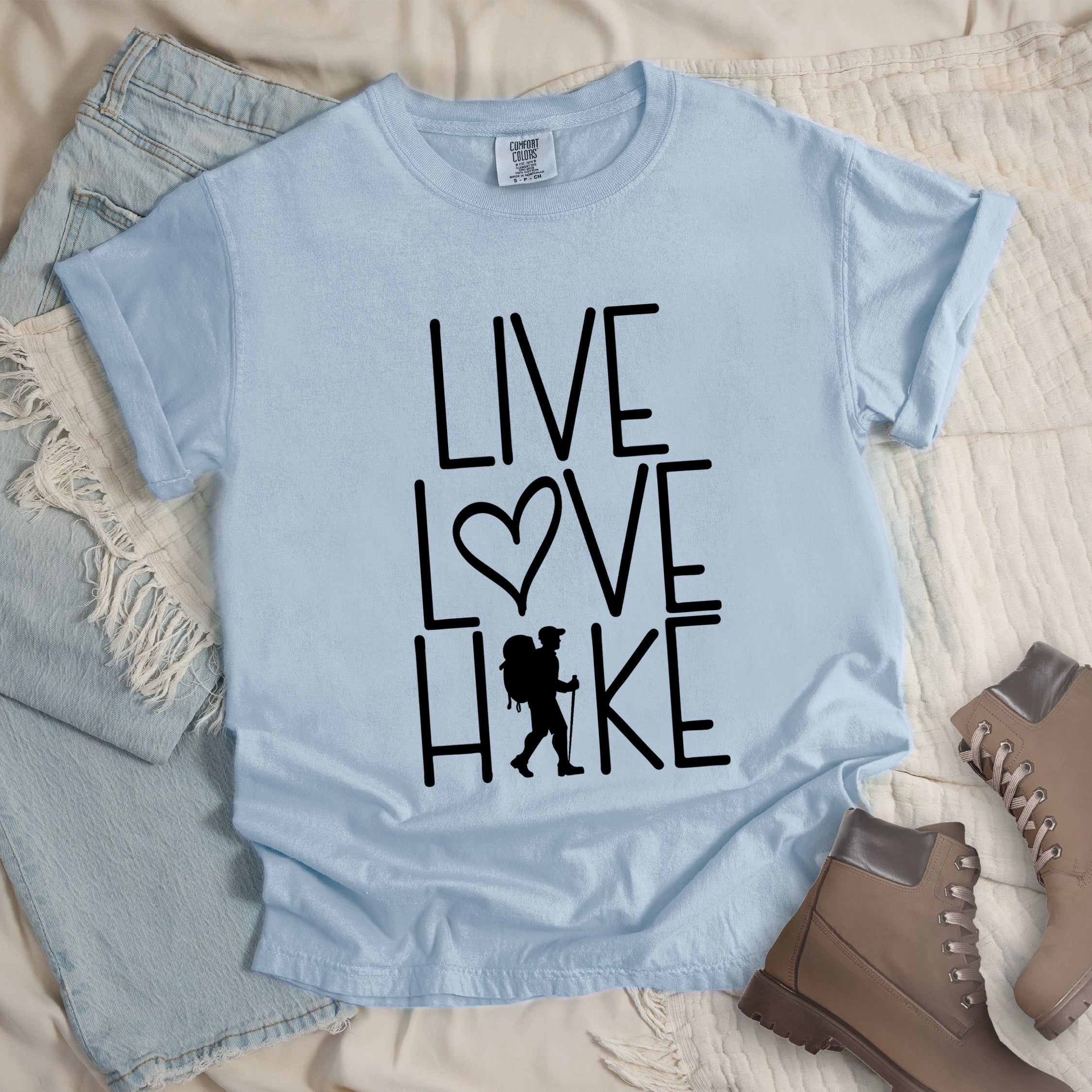 Chambray (sky blue) colored t-shirt with black text reading 'Live Love Hike' featuring a heart symbol and a male hiker silhouette with backpack, styled with light wash denim jeans and brown hiking boots on white bedding.