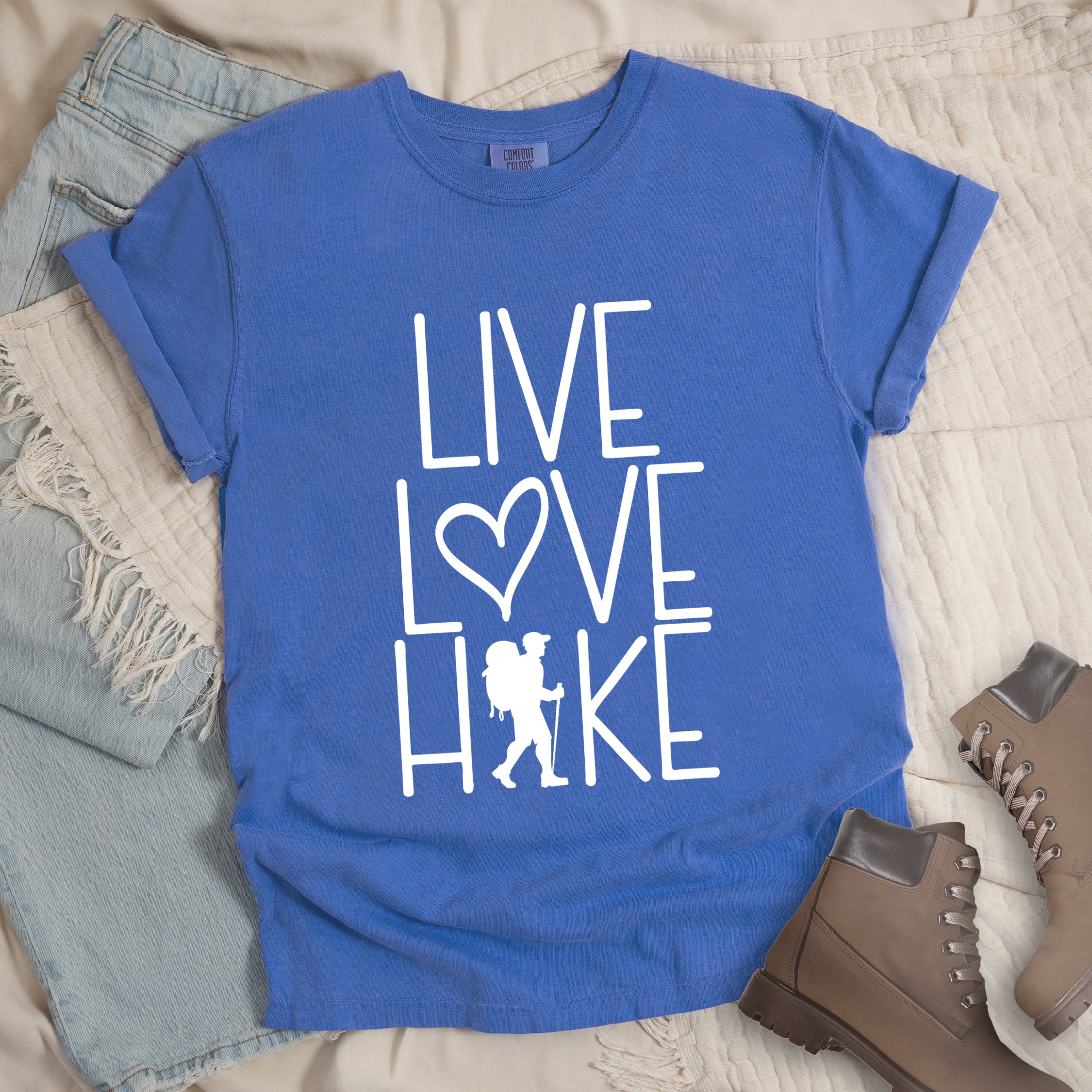 Flo Blue colored t-shirt with white text reading 'Live Love Hike' featuring a heart symbol and a male hiker silhouette with backpack, styled with light wash denim jeans and brown hiking boots on white bedding.