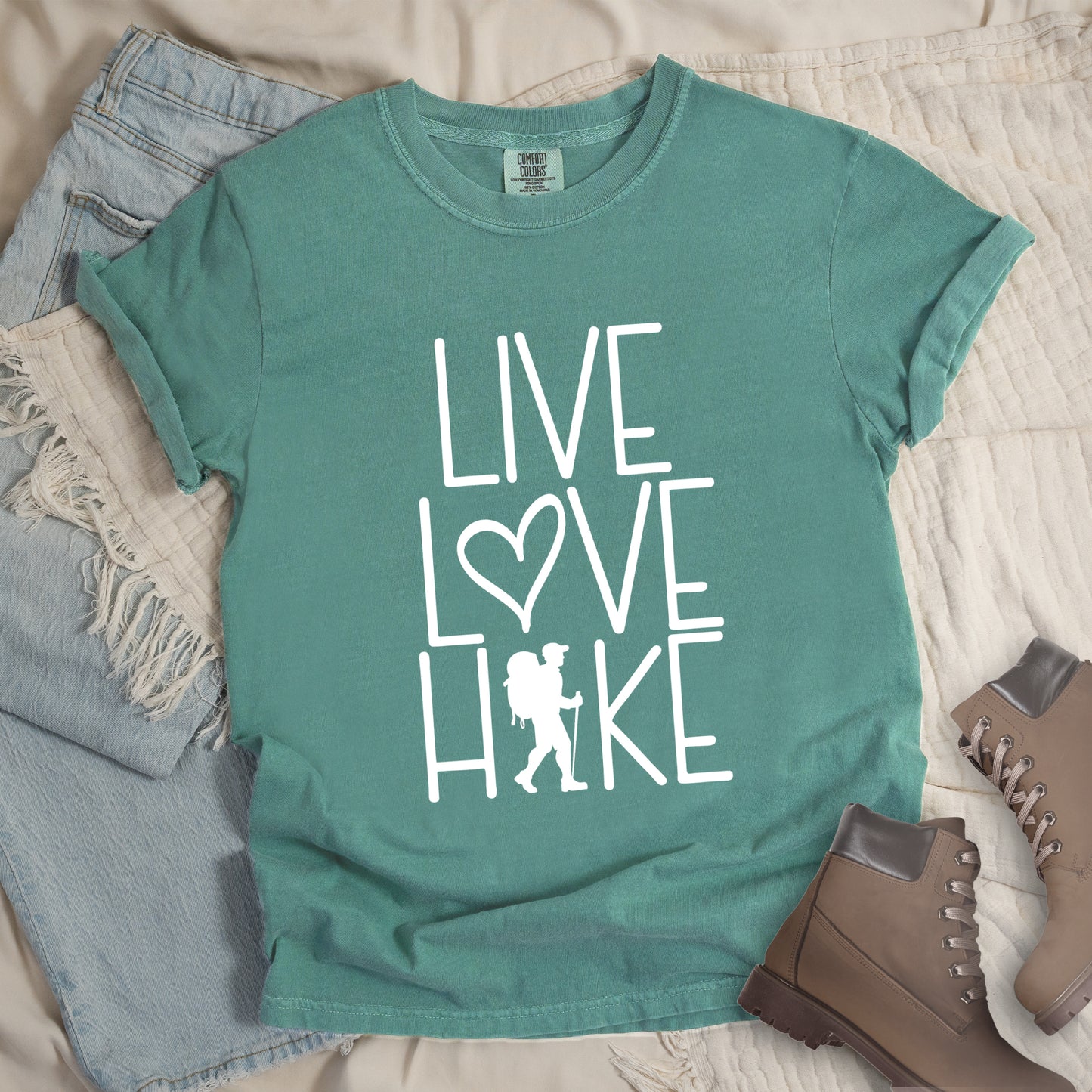 Light Green colored t-shirt with white text reading 'Live Love Hike' featuring a heart symbol and a male hiker silhouette with backpack, styled with light wash denim jeans and brown hiking boots on white bedding.