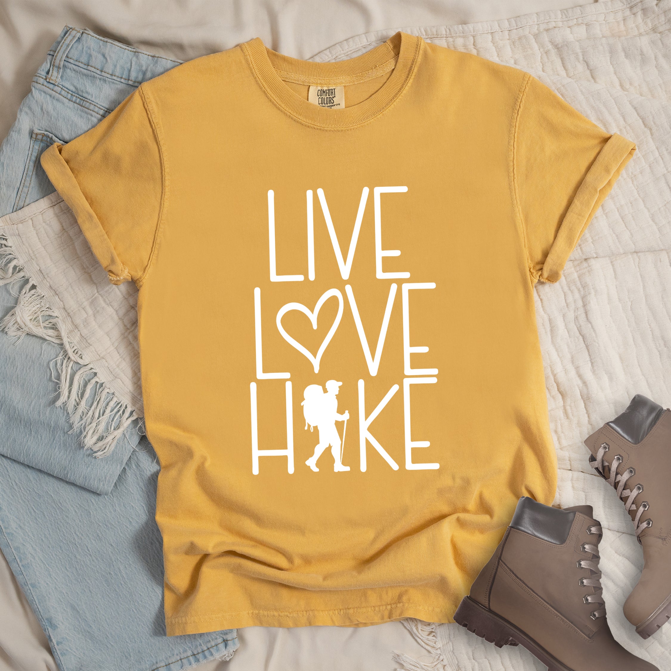 Mustard colored t-shirt with white text reading 'Live Love Hike' featuring a heart symbol and a male hiker silhouette with backpack, styled with light wash denim jeans and brown hiking boots on white bedding.