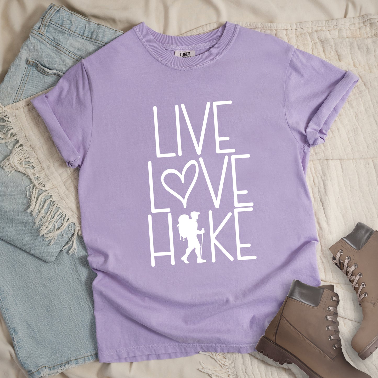 Orchid (lavender) colored t-shirt with white text reading 'Live Love Hike' featuring a heart symbol and a male hiker silhouette with backpack, styled with light wash denim jeans and brown hiking boots on white bedding.