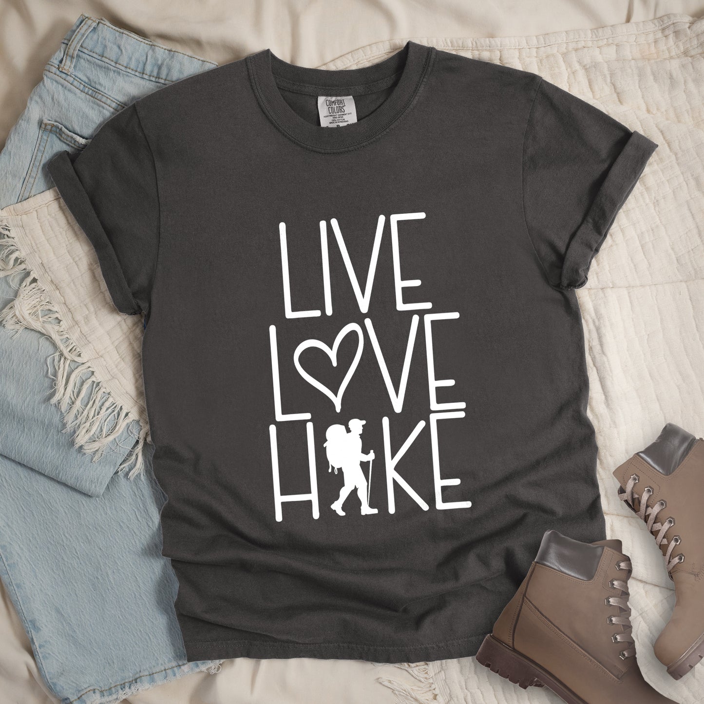 Pepper colored t-shirt with white text reading 'Live Love Hike' featuring a heart symbol and a male hiker silhouette with backpack, styled with light wash denim jeans and brown hiking boots on white bedding.