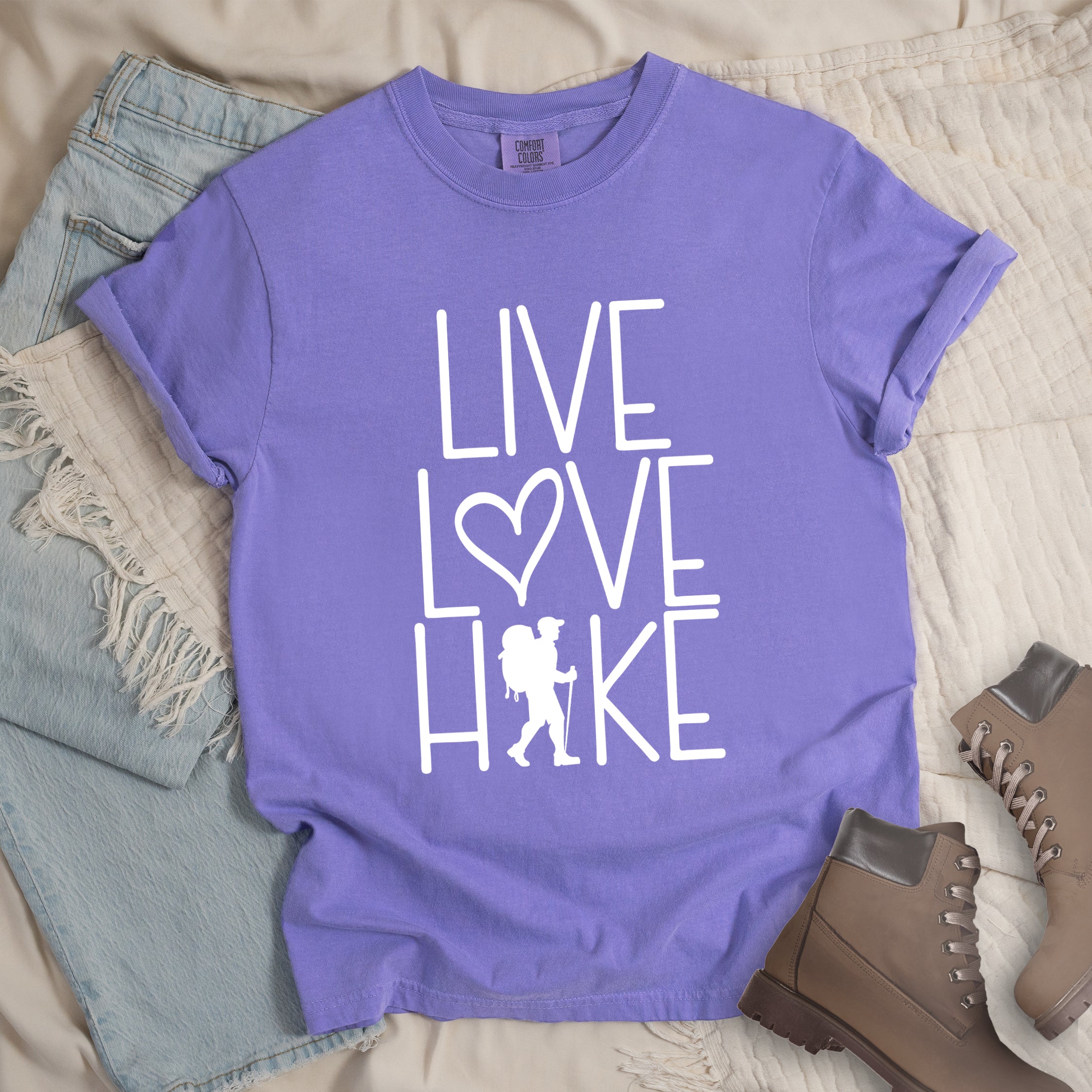 Violet colored t-shirt with white text reading 'Live Love Hike' featuring a heart symbol and a male hiker silhouette with backpack, styled with light wash denim jeans and brown hiking boots on white bedding.