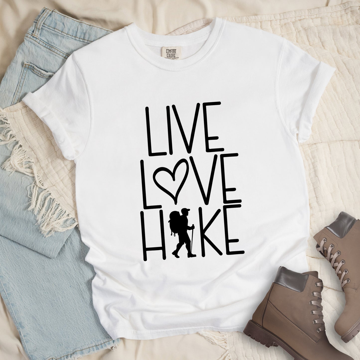 White colored t-shirt with black text reading 'Live Love Hike' featuring a heart symbol and a male hiker silhouette with backpack, styled with light wash denim jeans and brown hiking boots on white bedding.