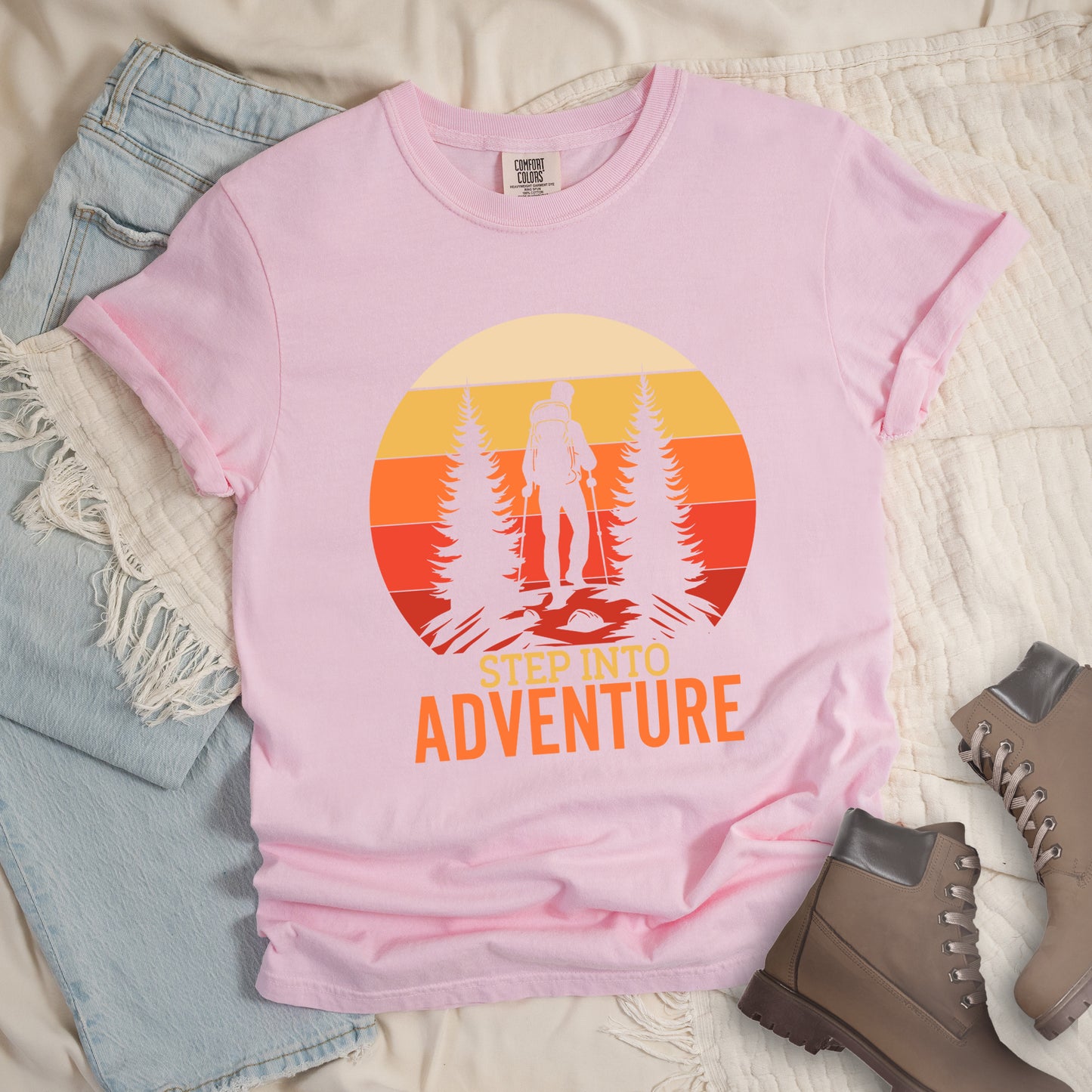 Blossom (Light Pink) graphic t-shirt with 'Step Into Adventure' text and vintage sunset design featuring hiker silhouette among pine trees, styled with light wash jeans and brown hiking boots