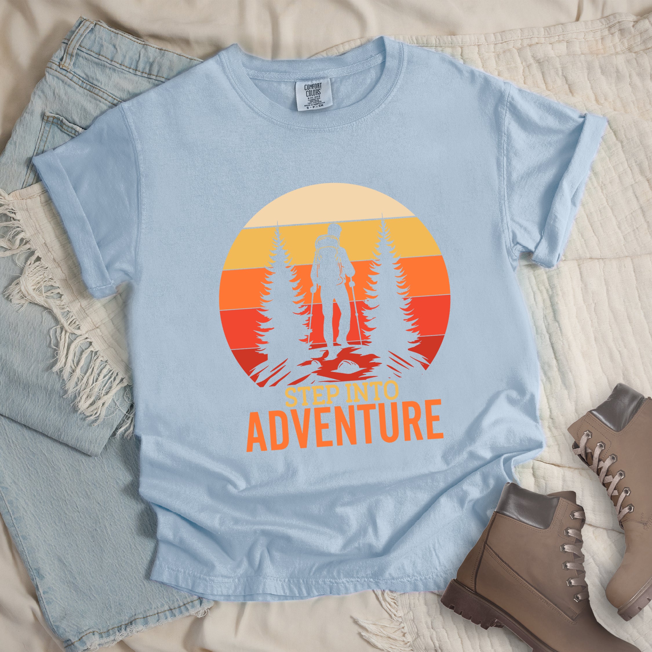 Chambray (Light Blue) graphic t-shirt with 'Step Into Adventure' text and vintage sunset design featuring hiker silhouette among pine trees, styled with light wash jeans and brown hiking boots