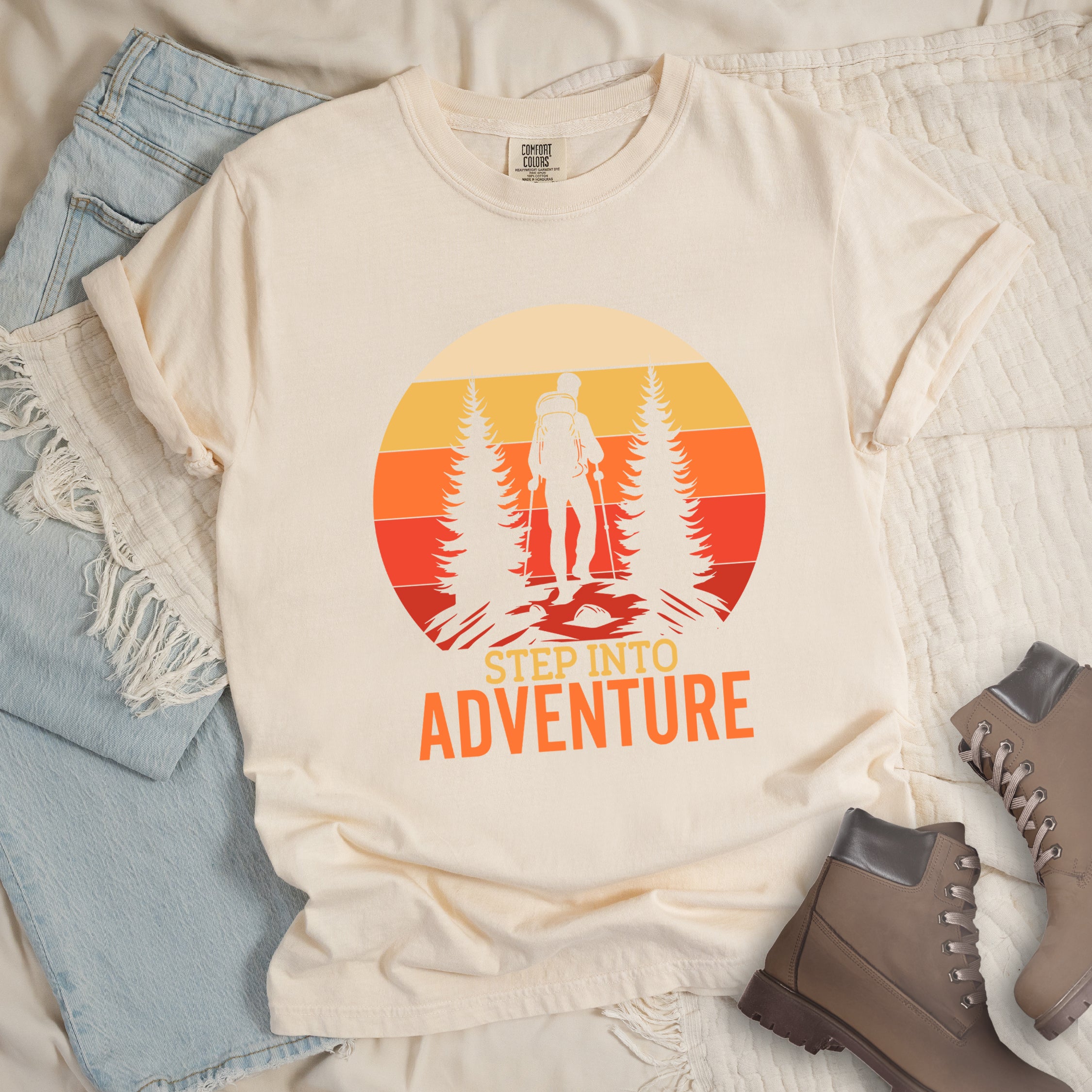 Ivory (Cream White) graphic t-shirt with 'Step Into Adventure' text and vintage sunset design featuring hiker silhouette among pine trees, styled with light wash jeans and brown hiking boots