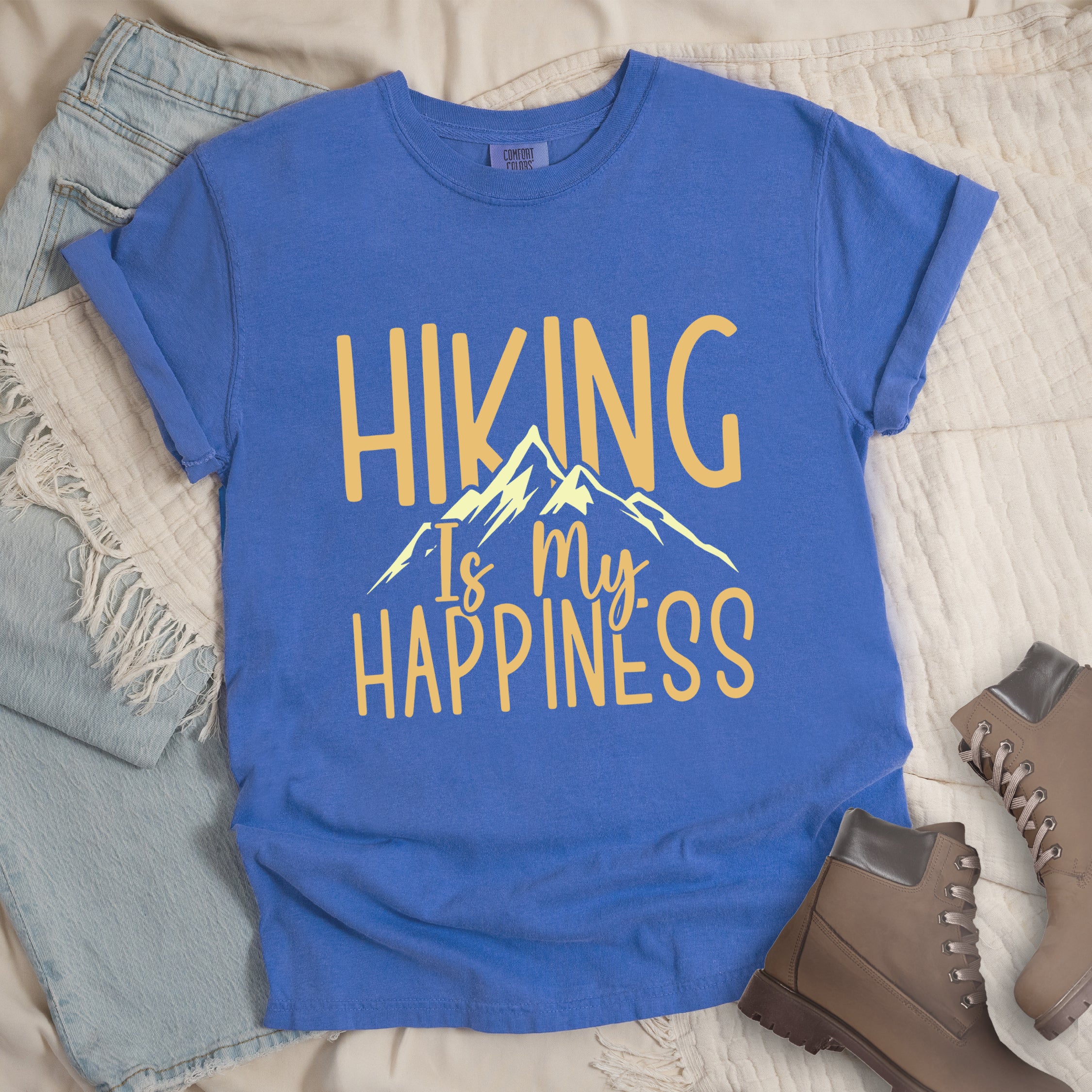 Flo Blue (Bright Blue) graphic t-shirt with 'Hiking Is My Happiness' text in yellow and cream mountain design, styled with light wash jeans and brown hiking boots