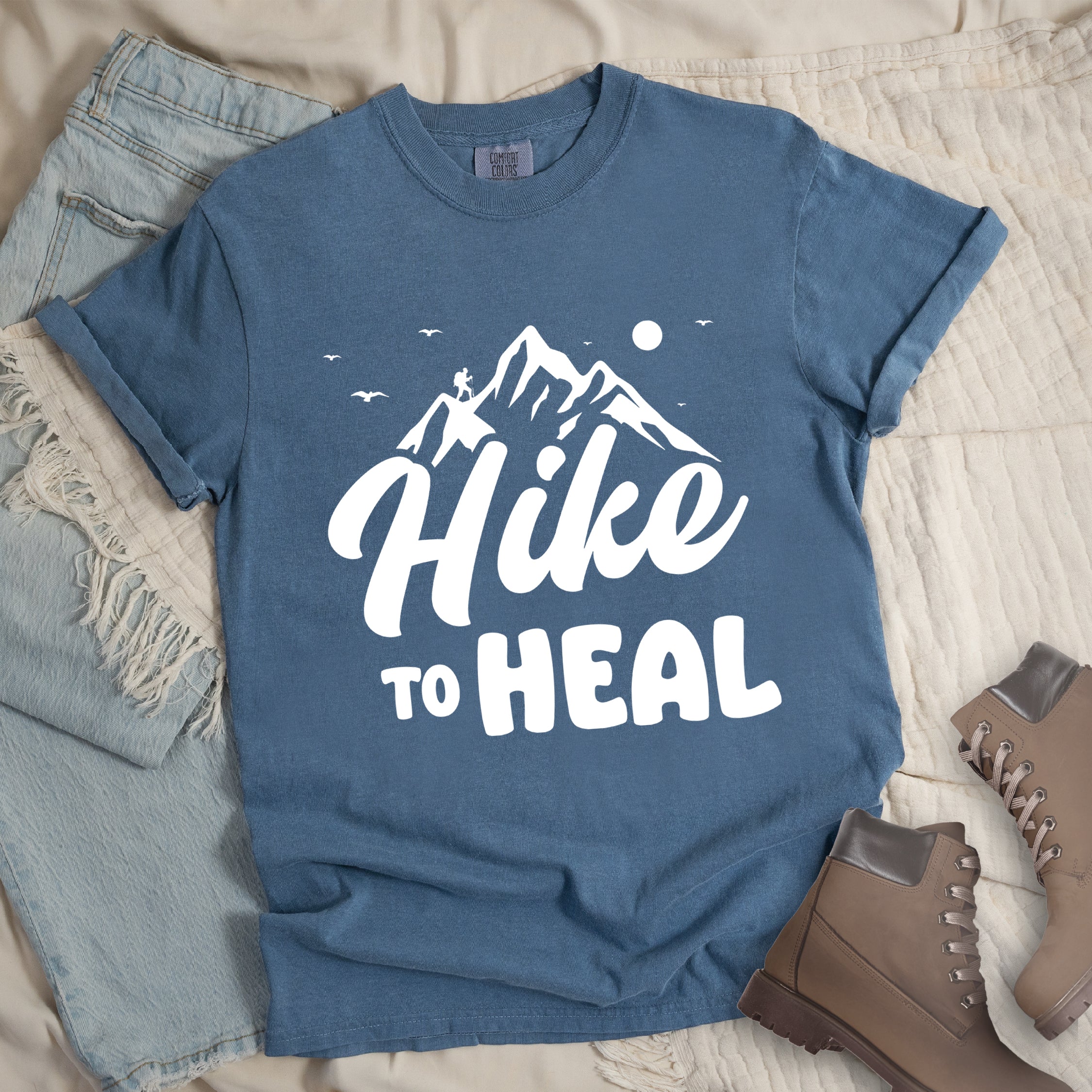 Blue Jean (Denim Blue) graphic t-shirt with 'Hike to Heal' text in white with mountain and bird design, styled with light wash jeans and brown hiking boots