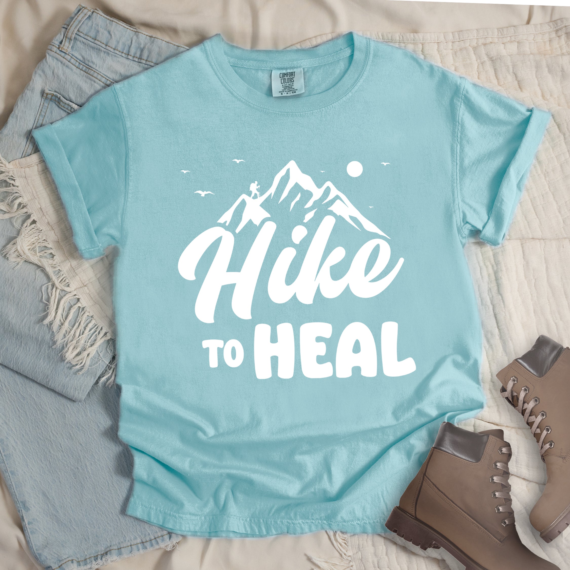 Chalky Mint (Pastel Blue) graphic t-shirt with 'Hike to Heal' text in white with mountain and bird design, styled with light wash jeans and brown hiking boots
