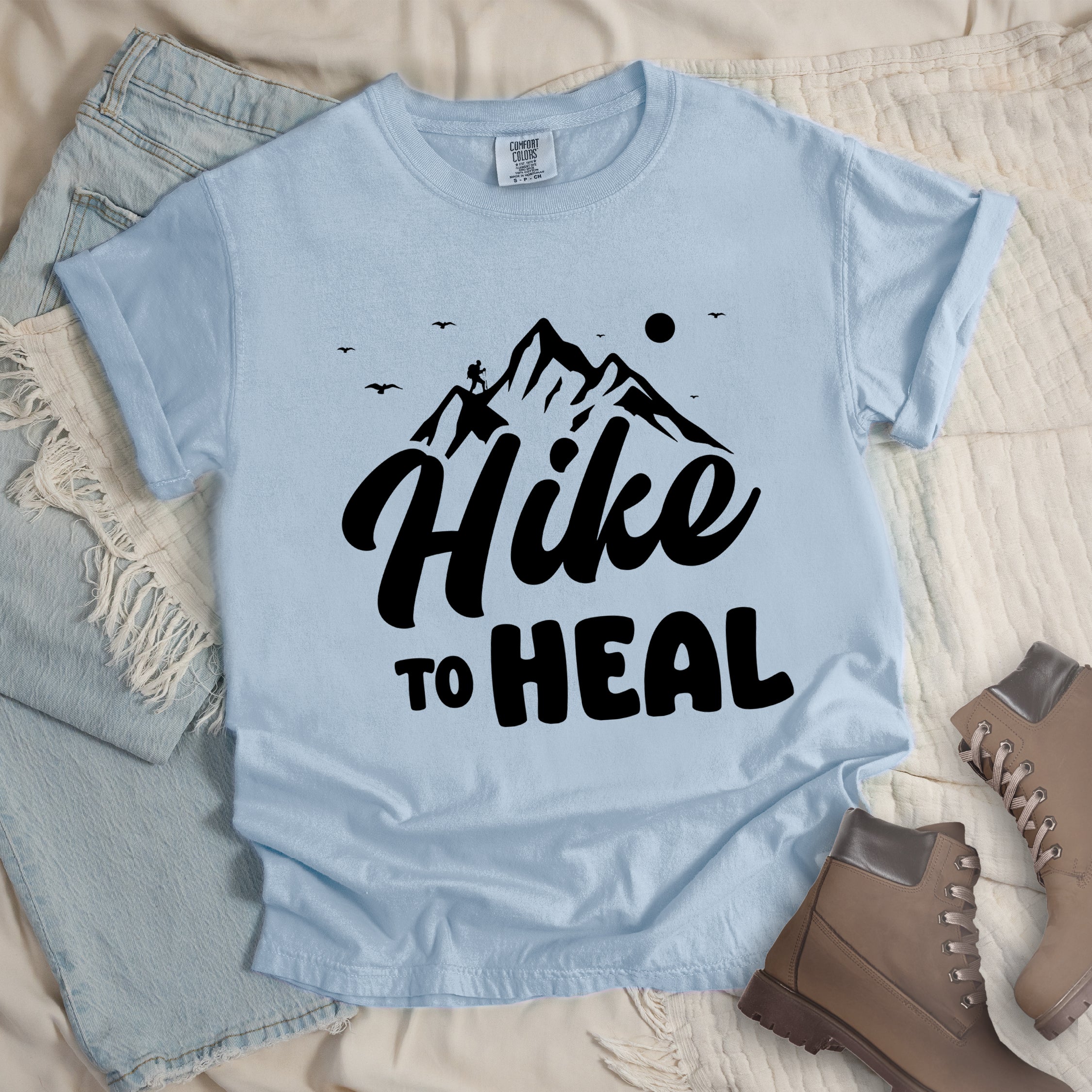 Chambray (Light Blue) graphic t-shirt with 'Hike to Heal' text in white with mountain and bird design, styled with light wash jeans and brown hiking boots