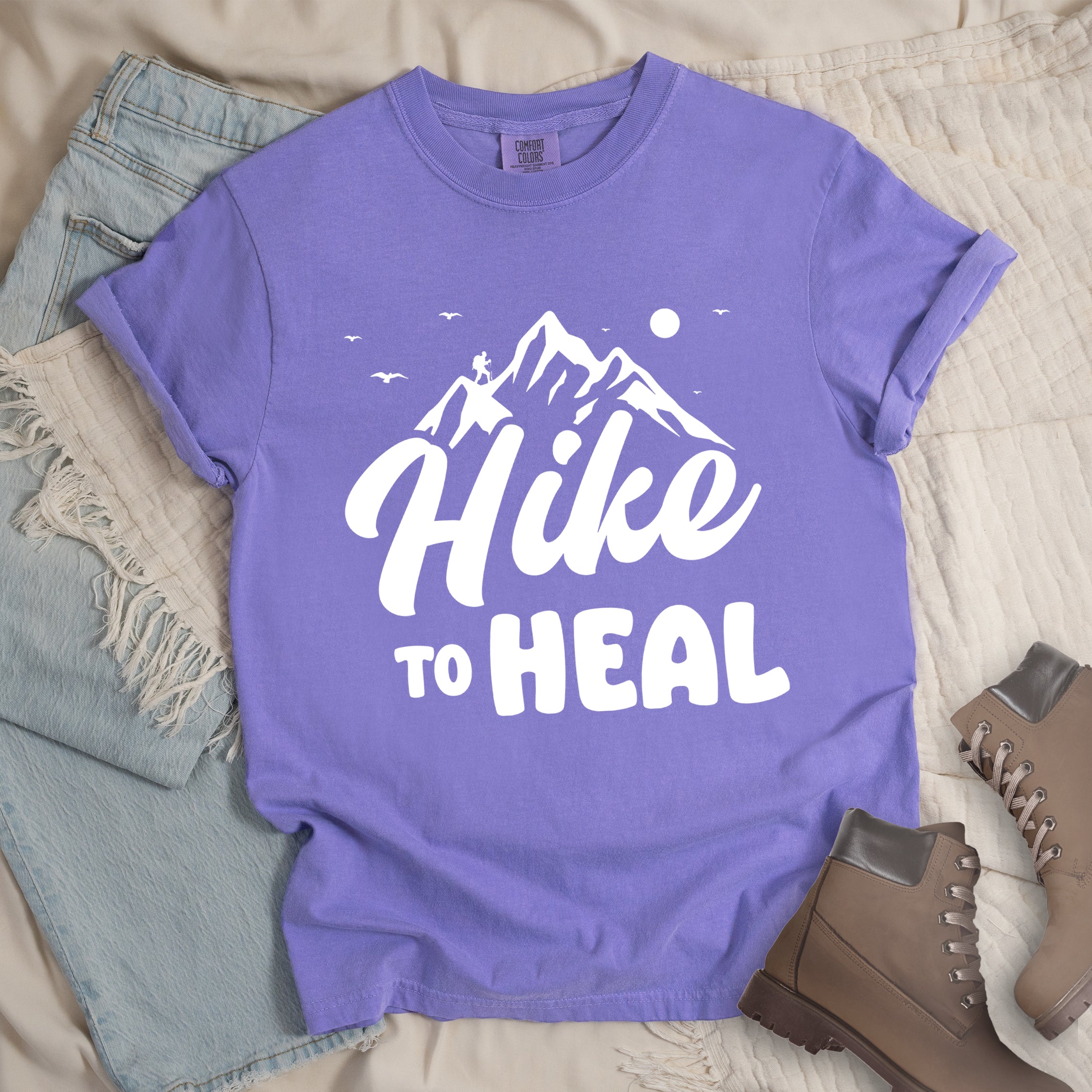 Violet graphic t-shirt with 'Hike to Heal' text in white with mountain and bird design, styled with light wash jeans and brown hiking boots