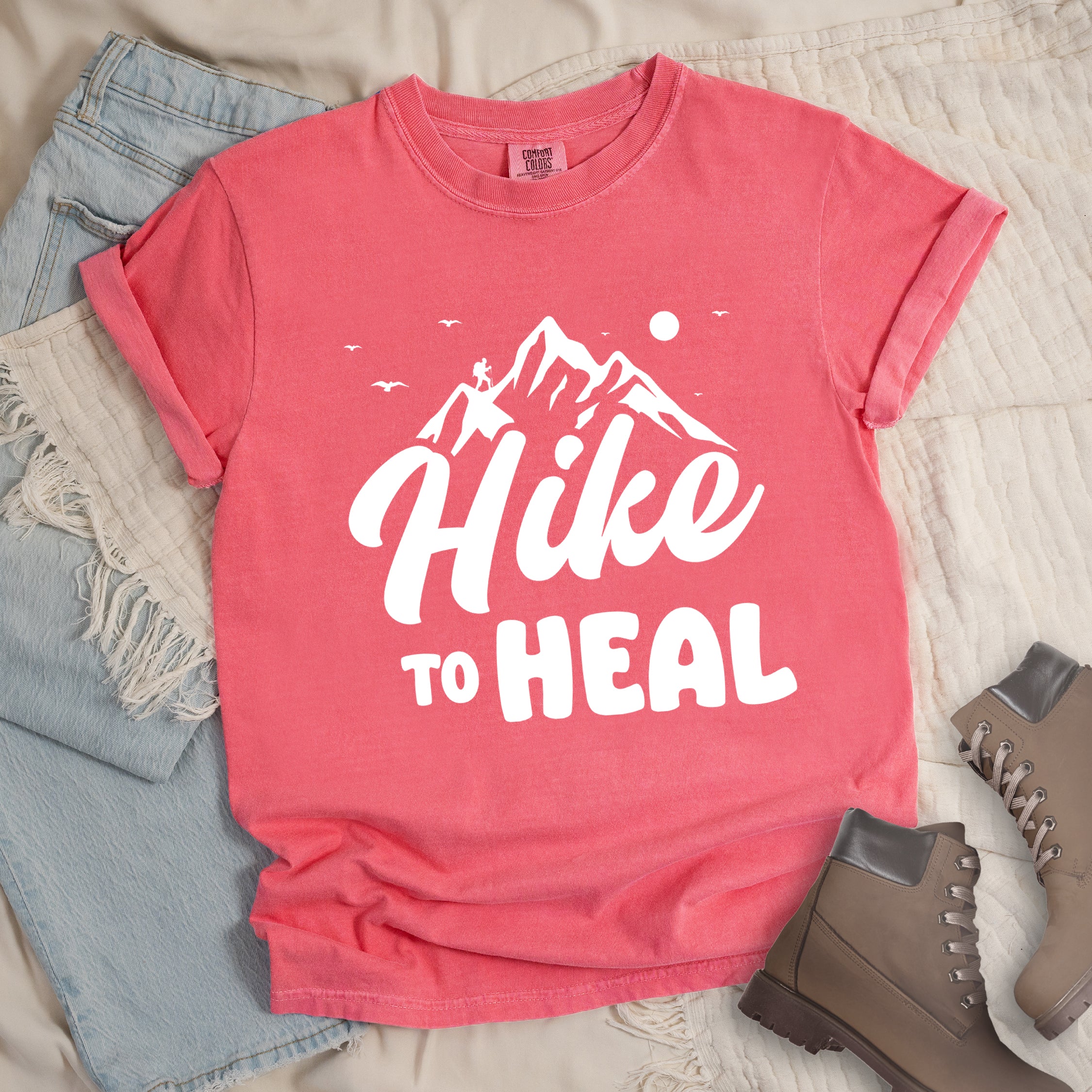 Watermelon (Light Coral) graphic t-shirt with 'Hike to Heal' text in white with mountain and bird design, styled with light wash jeans and brown hiking boots