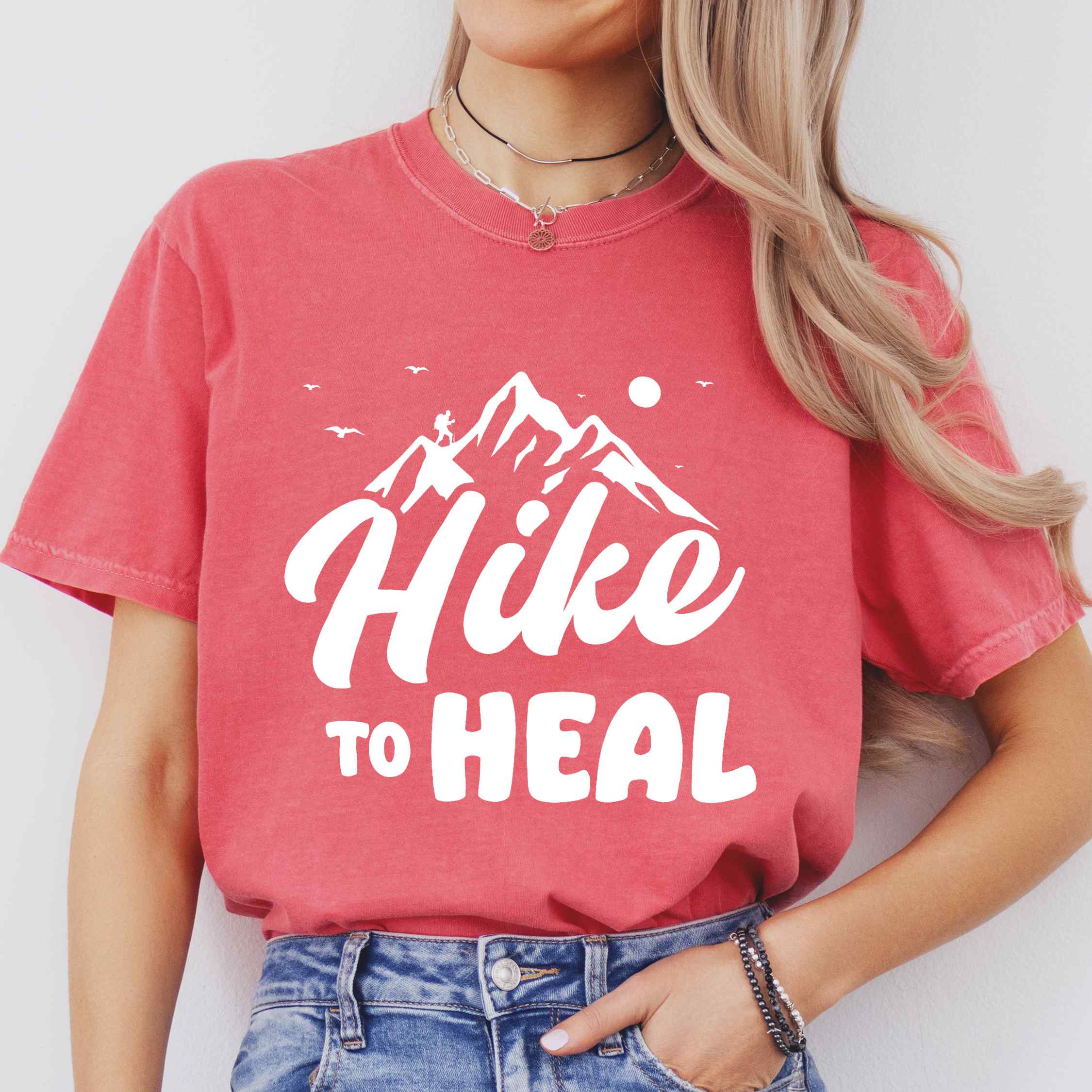 Woman wearing watermelon "Hike to Heal" colored statement tee with bold typography, mountain, sunrise, perfect for adventure driven individuals.